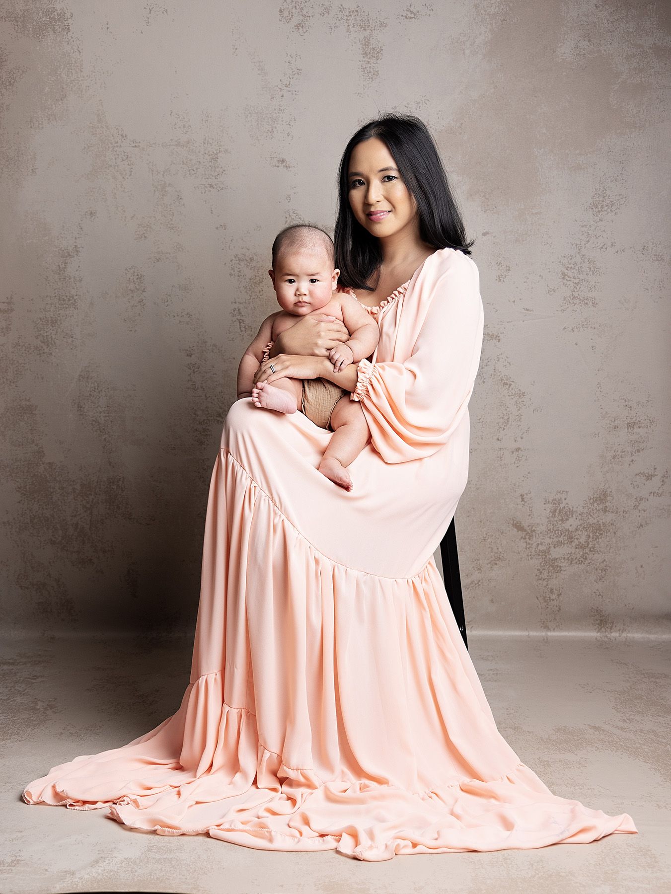 Portrait of parent and baby in matching peach-colored outfits against textured gray backdrop during professional photo session.