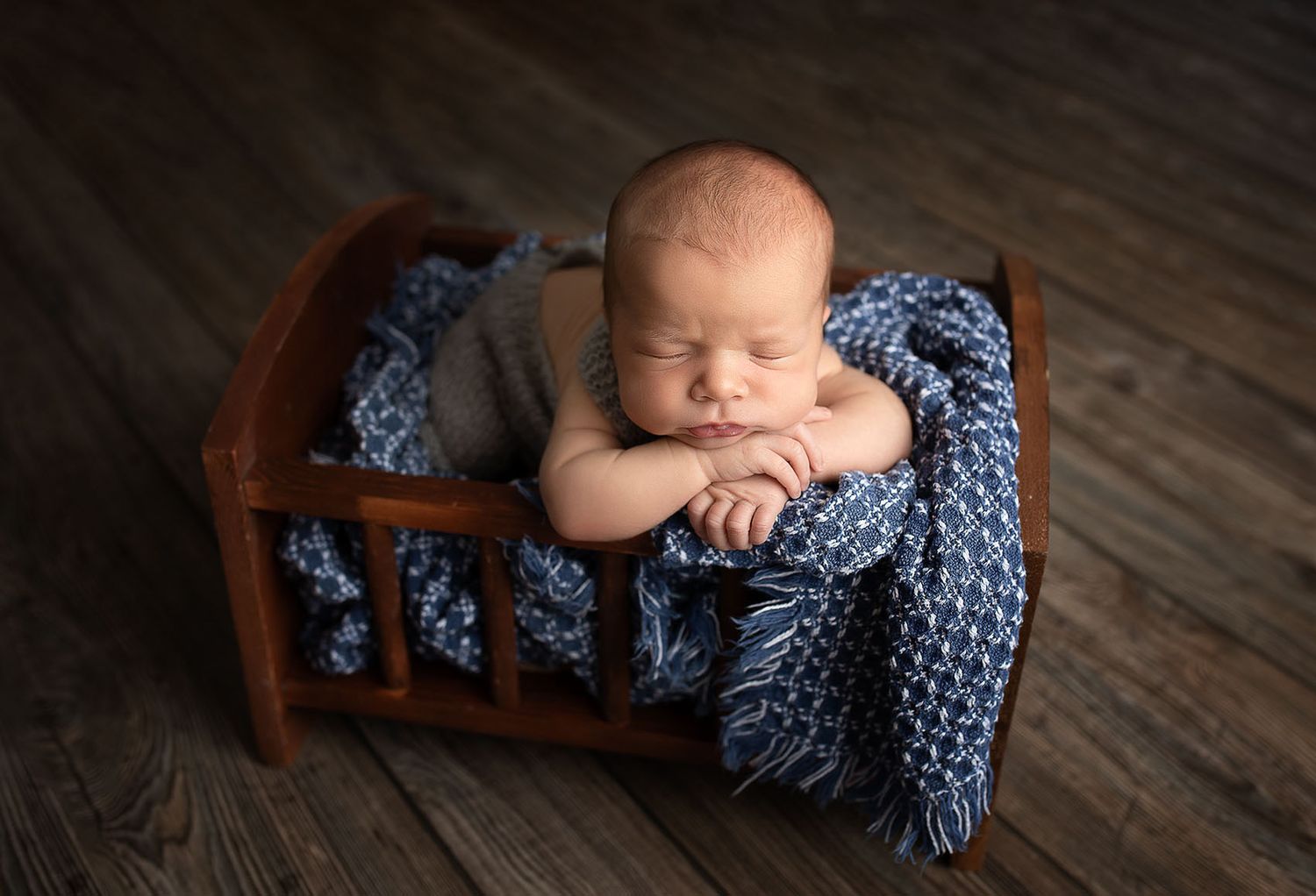 Sleeping newborn rests peacefully in small wooden bed with blue knit blanket on dark wooden floor.