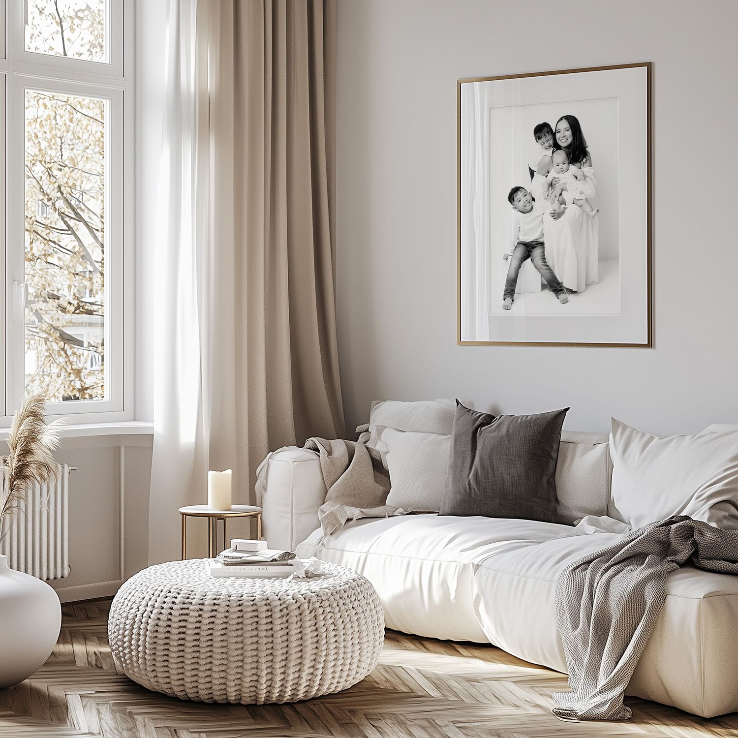 Cozy Scandinavian living room with white sofa, knit pouf, framed wall art, and natural light from large window.
