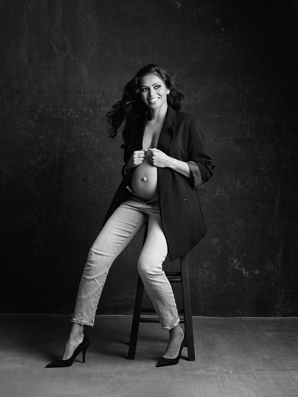Black and white maternity portrait showing a pregnant woman in a blazer and pants posing elegantly on a dark background.