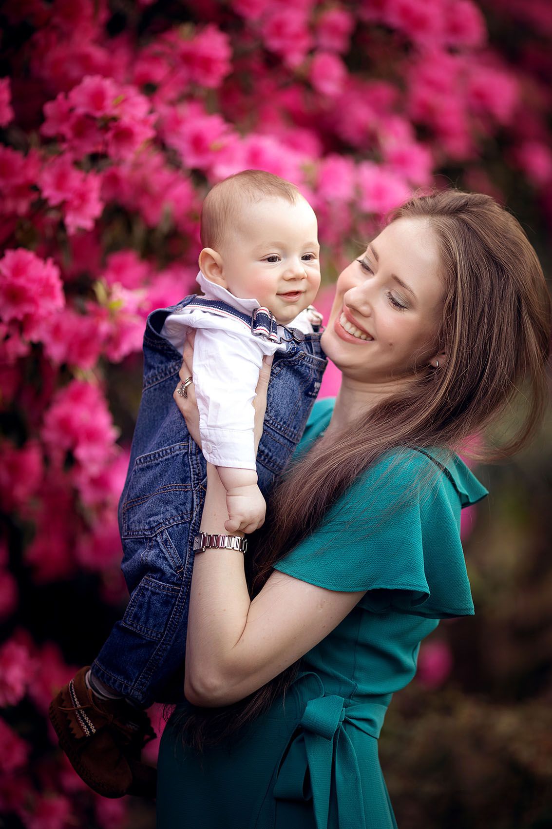 A tender moment captured against vibrant pink azalea blossoms with a baby in denim overalls and woman in emerald green dress.
