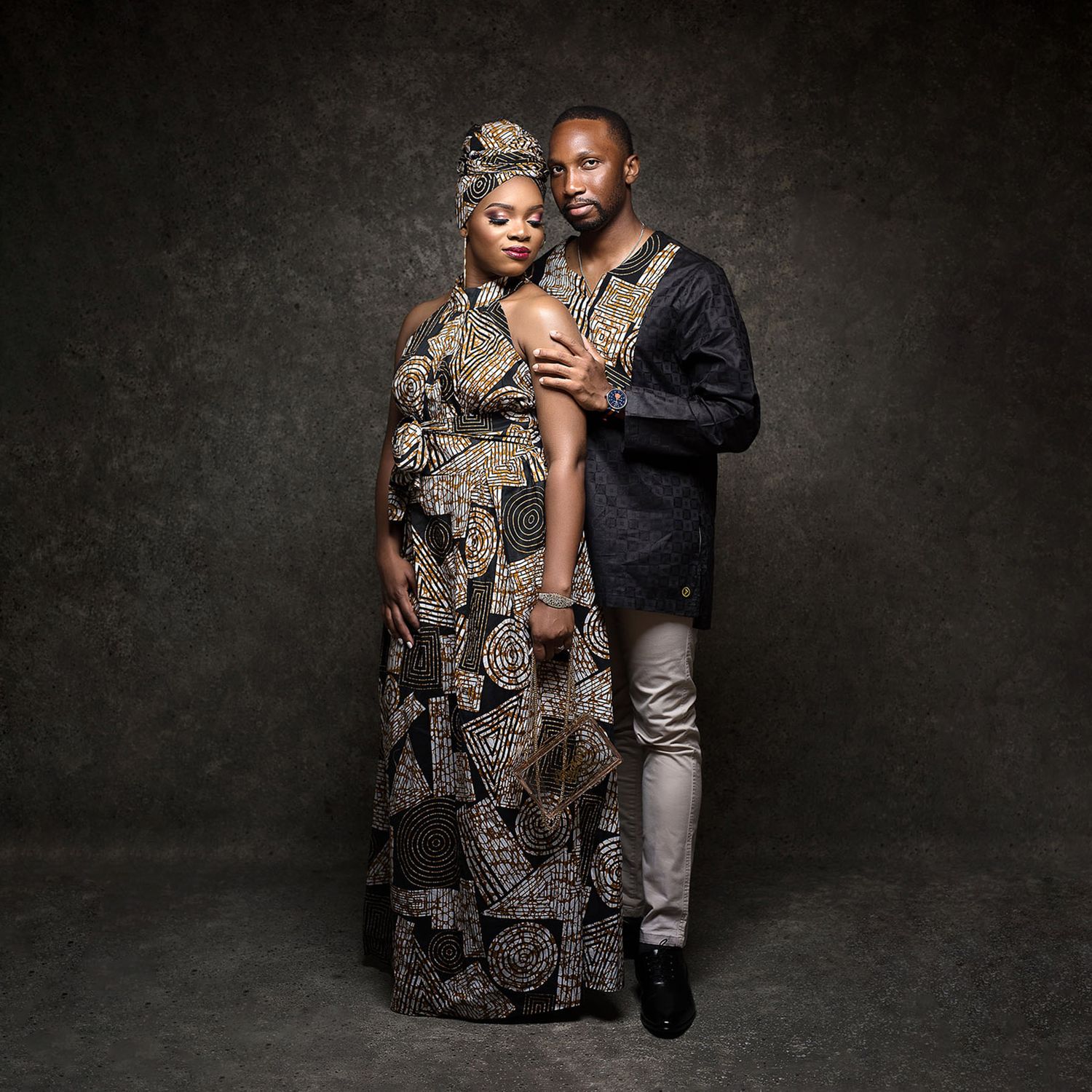 A couple in elegant attire pose together against a dark dramatic backdrop in an artistic portrait.