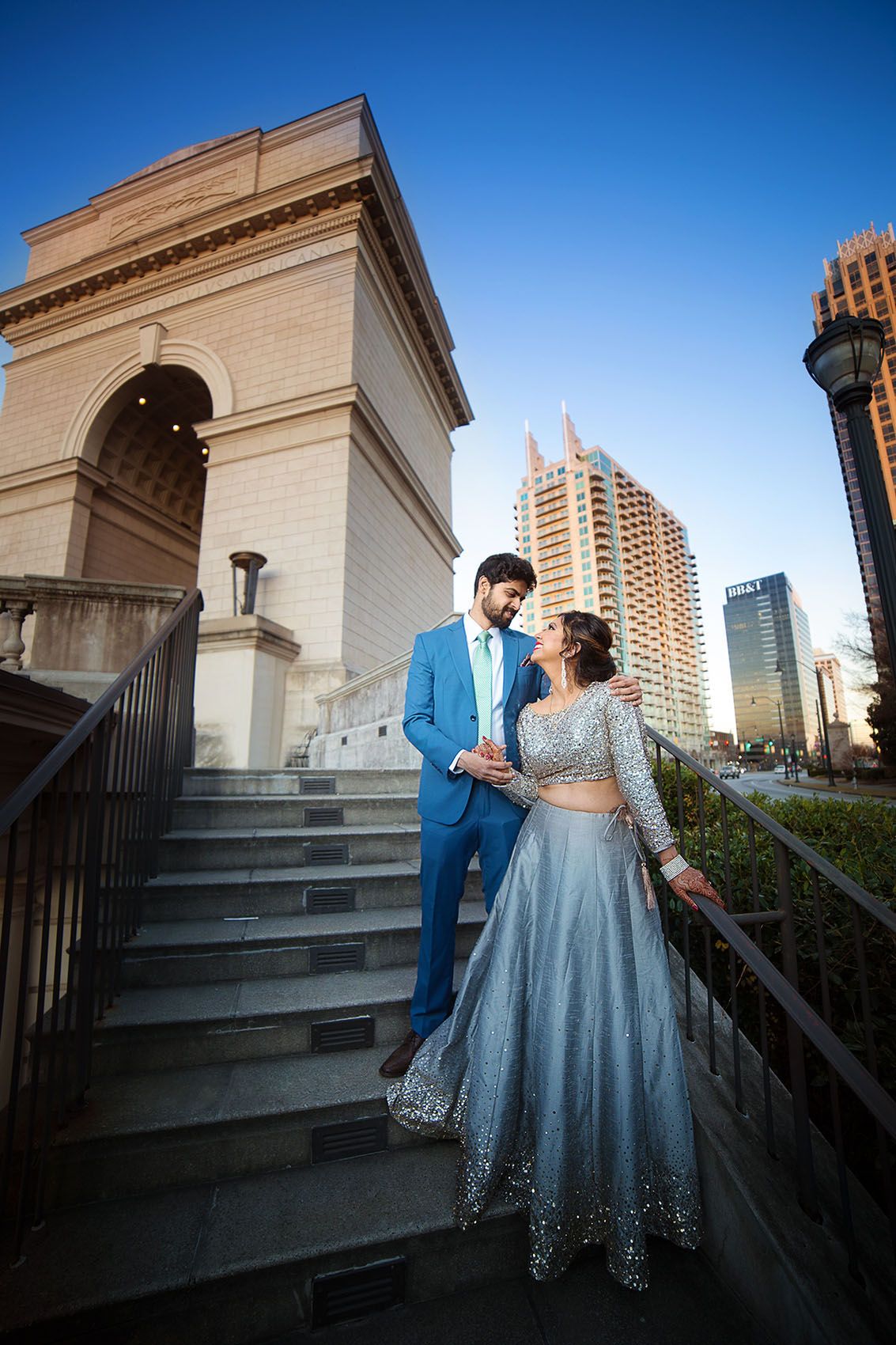 A couple in formal blue attire stand on stone steps against an urban backdrop with classical architecture and skyscrapers.