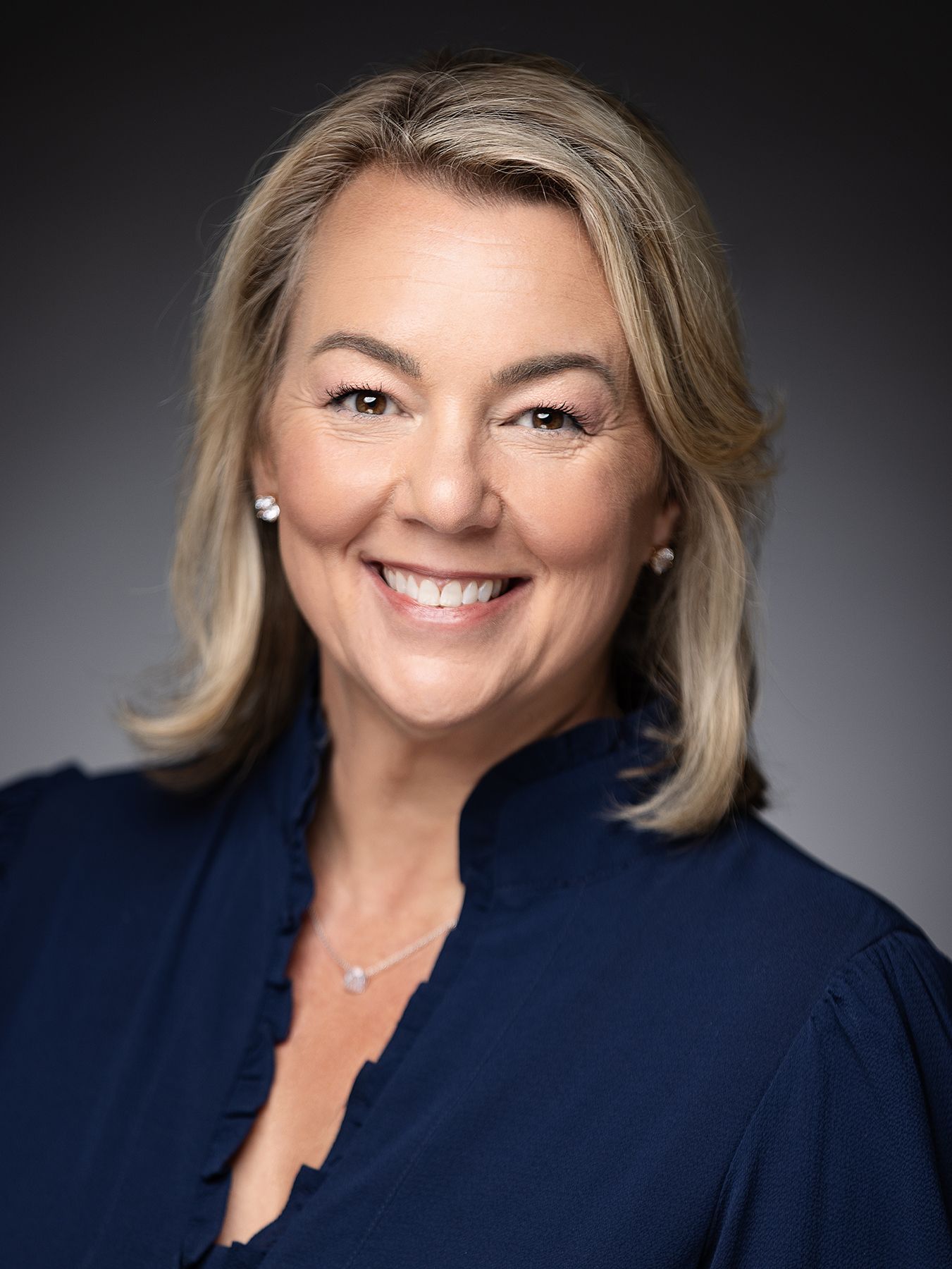 Professional headshot of a person in navy blazer with shoulder-length blonde hair against a dark gray background.