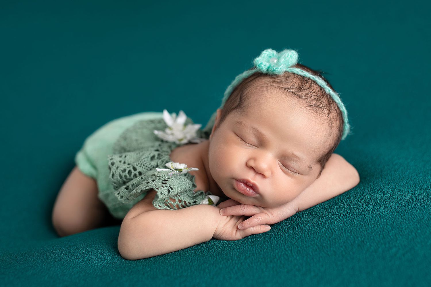 Peaceful sleeping newborn wearing mint green lace outfit lies on teal backdrop during professional photo session.