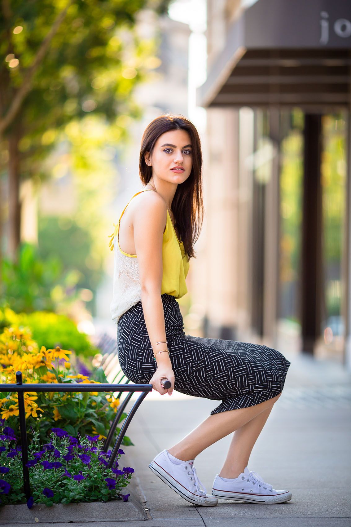 A person in a yellow top and patterned pants sits on a railing near colorful flowers on an urban sidewalk.