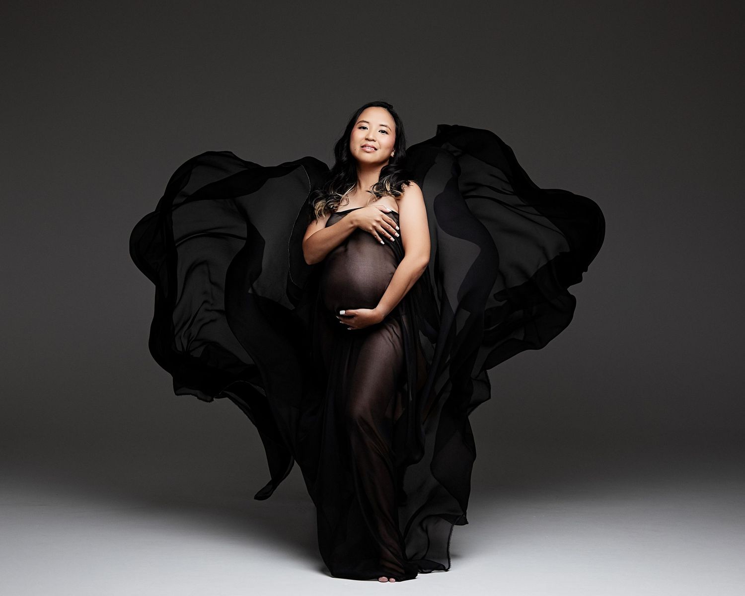 A silhouetted figure stands gracefully in flowing black fabric against a dark background during a dramatic studio photoshoot.