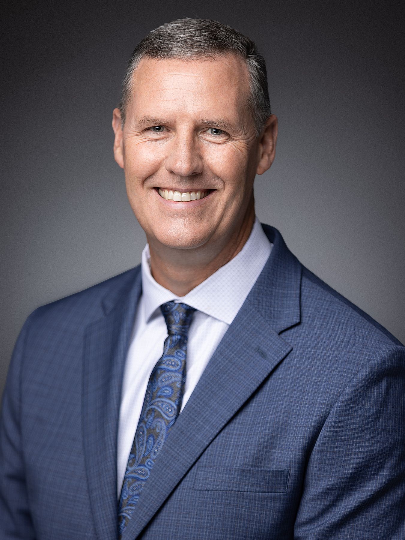 Professional business headshot of person in navy suit and blue patterned tie against gradient background.