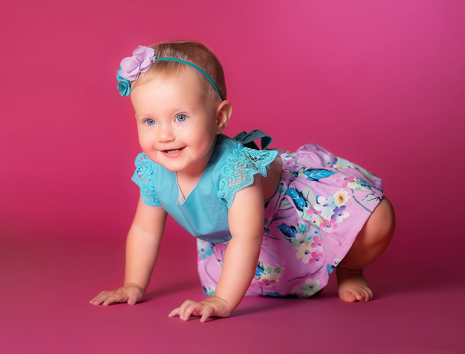 Baby wearing blue flutter sleeve top and pink floral romper crawls on pink studio backdrop.