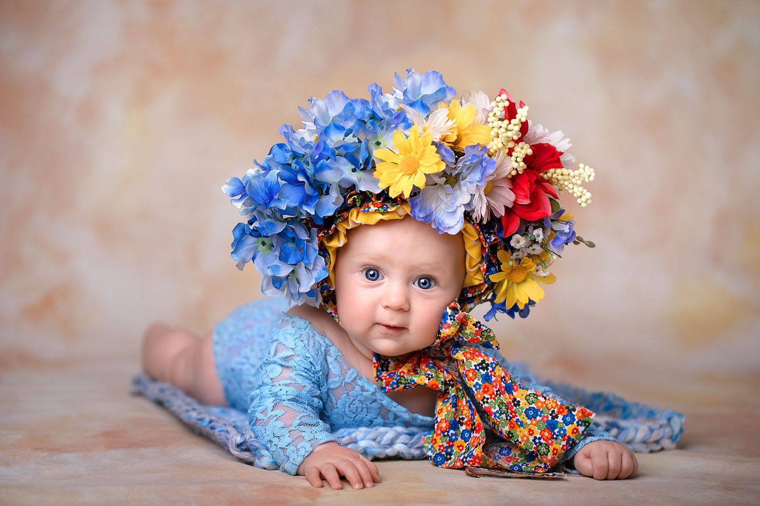 Baby wearing a floral crown made of blue, yellow and red flowers lies on a tan backdrop.