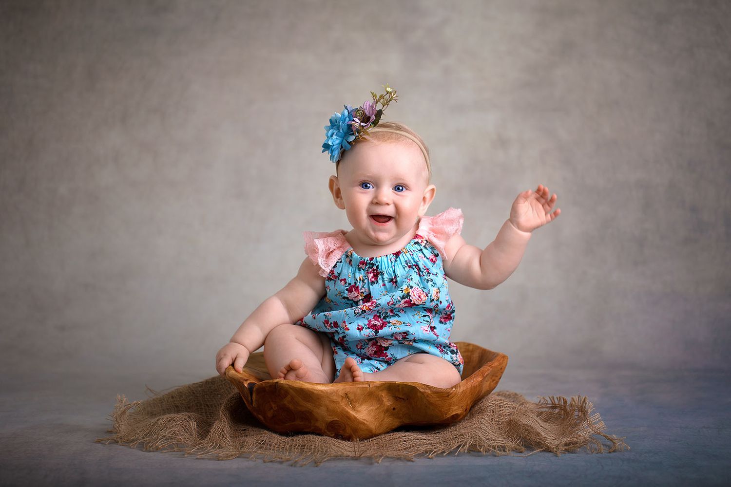 Happy infant in floral blue outfit sits in wooden bowl on burlap against gray backdrop, wearing hair bow.