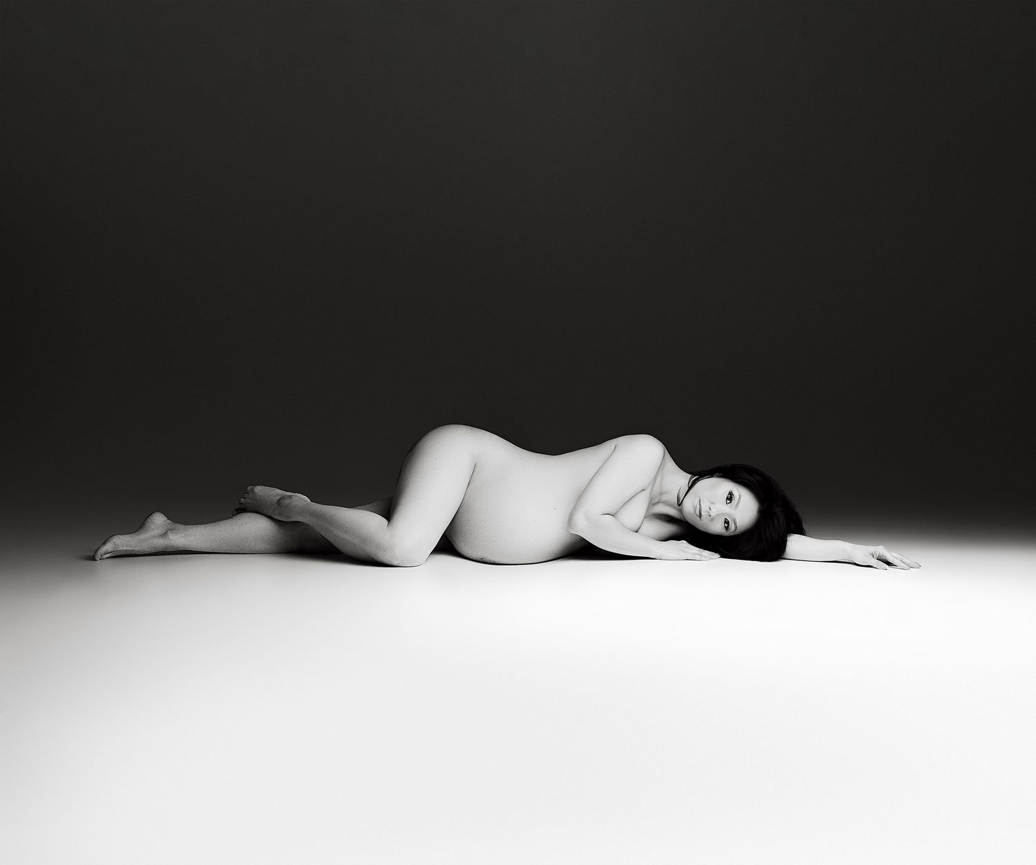 Artistic black and white nude photography showing reclining figure in minimalist setting against dark background.