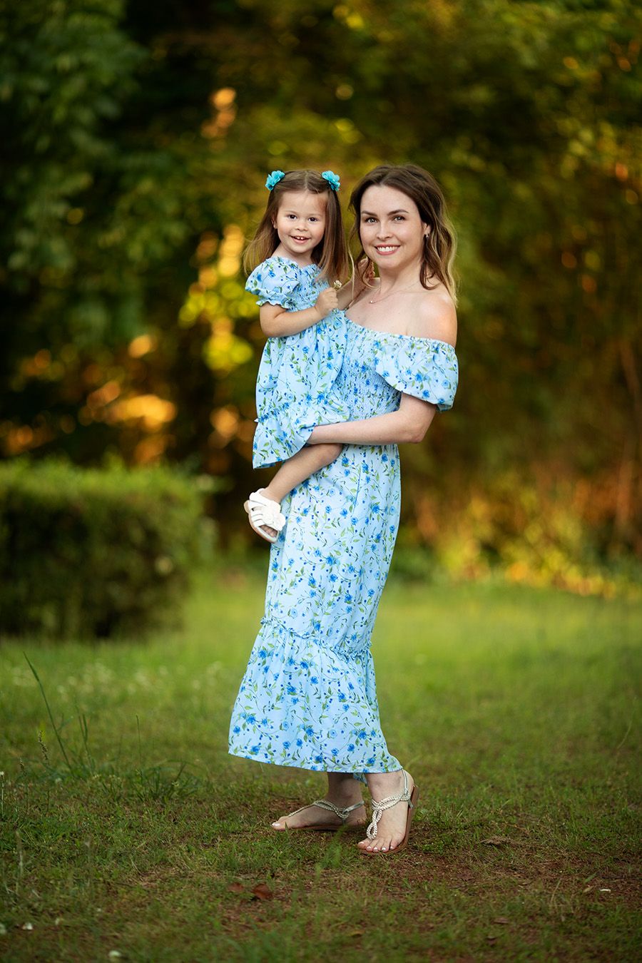 A mother and small child wearing matching light blue floral dresses pose together outdoors in a natural setting.