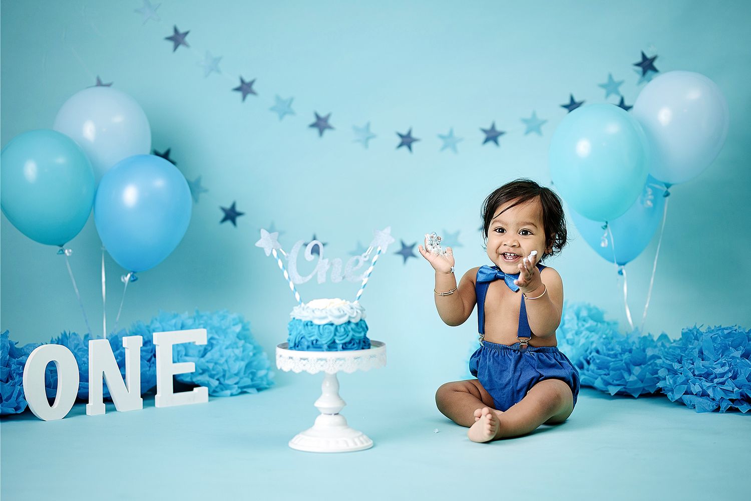 First birthday cake smash photo setup with blue balloons, silver stars, and 'ONE' letters against turquoise backdrop.