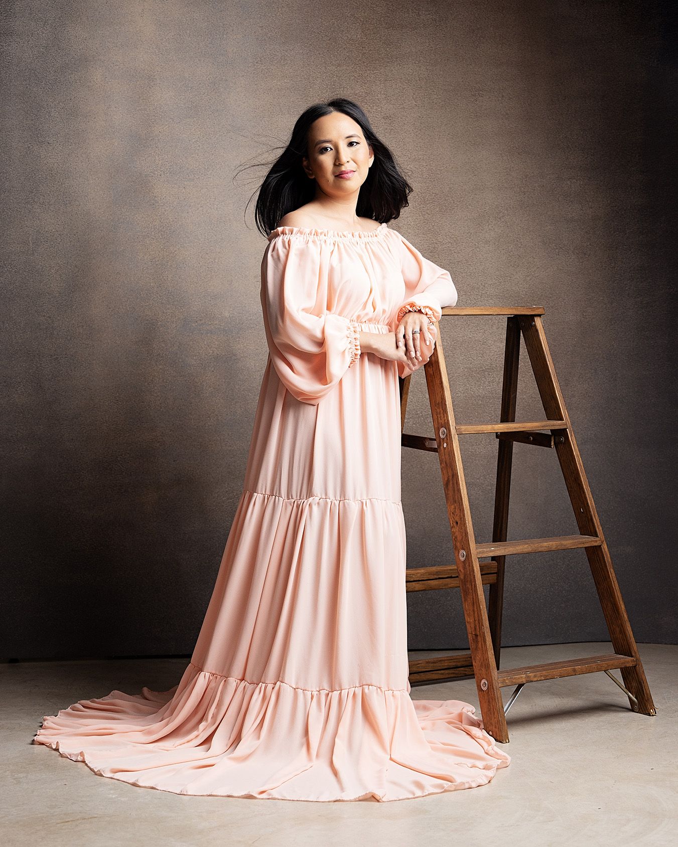 Woman in flowing pink gown poses elegantly in front of rustic ladder against dark gray studio backdrop.