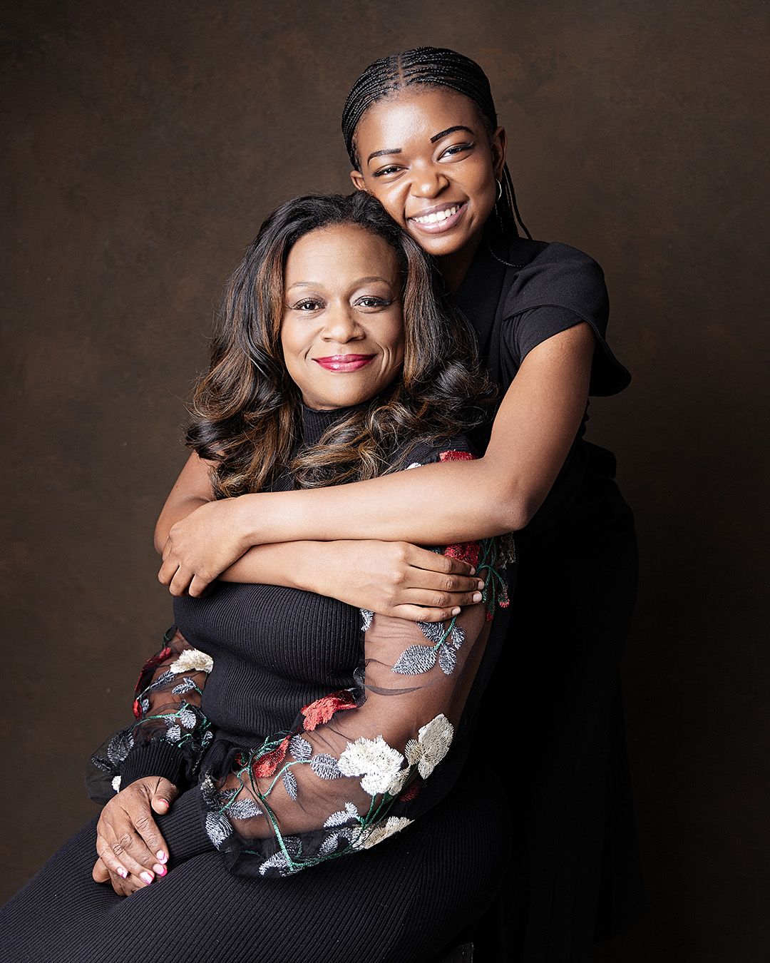 Two people in black outfits embrace while posing against a dark backdrop for a warm and intimate portrait.