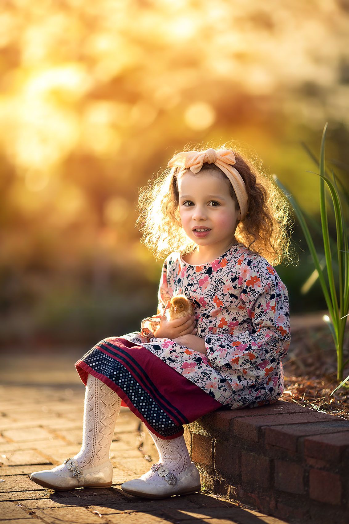 A child in a floral dress and white tights sits on brick steps during golden hour while holding a toy.