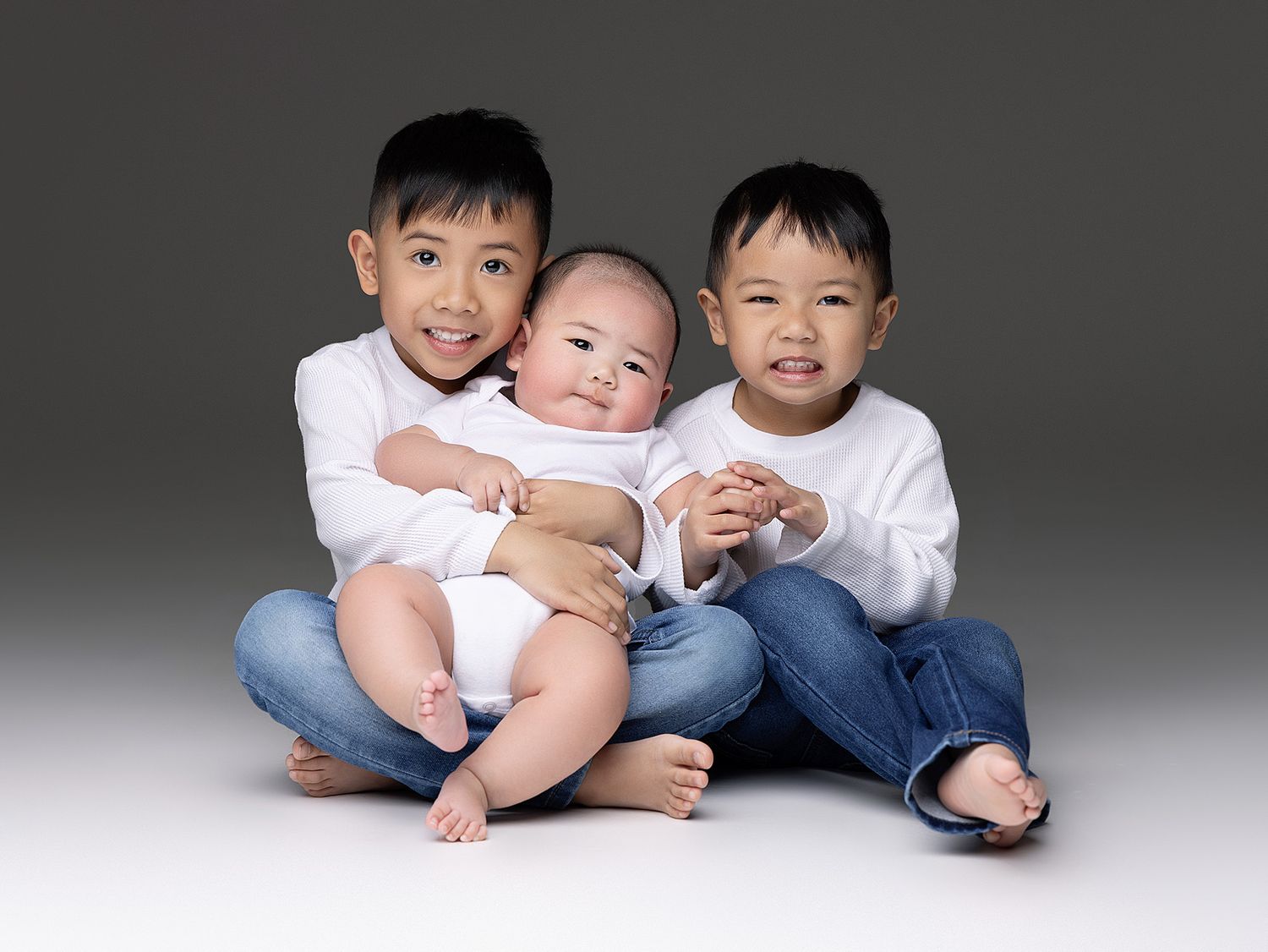 Three children in white shirts sit together on a gray background, with the youngest in the middle being embraced.