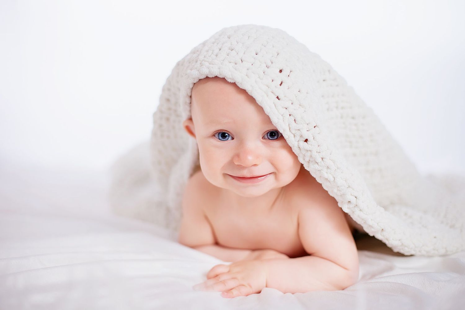 A happy baby lies under a white textured blanket against a light background, flashing a cheerful smile.