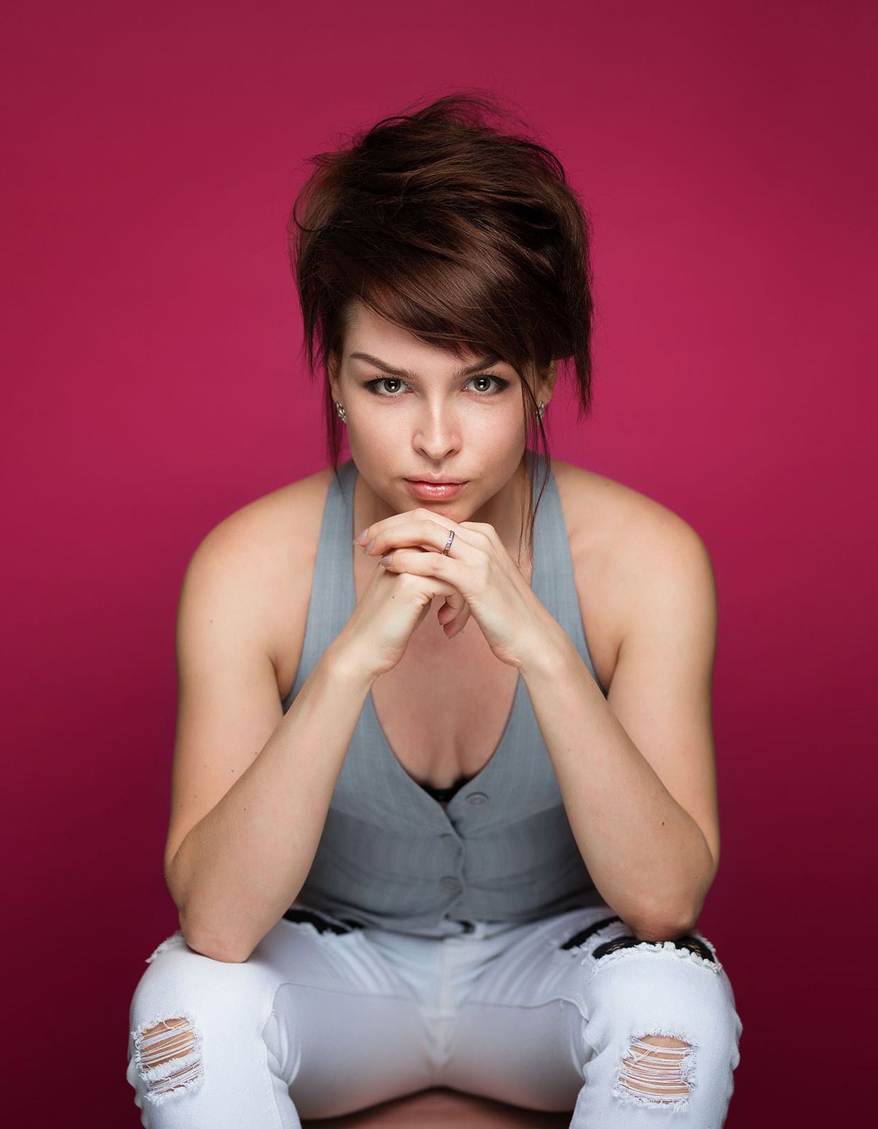 Model wearing a gray tank top and ripped white jeans poses thoughtfully against a burgundy background.