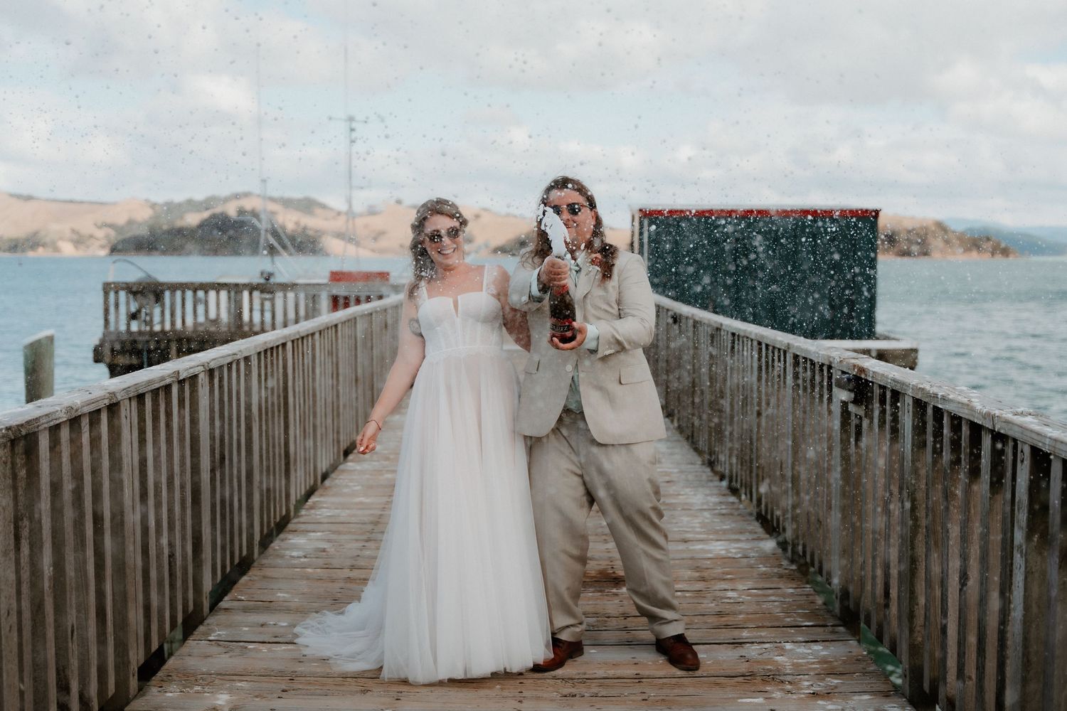 A couple stands together on a wooden pier overlooking the water and mountains on their wedding day in muted tones.