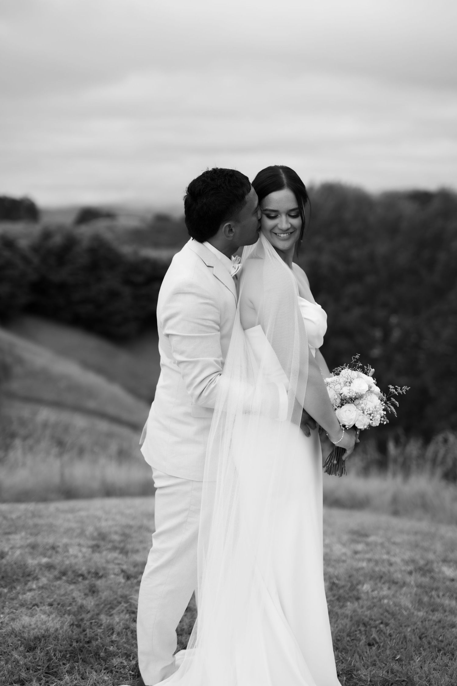 Black and white wedding photo of couple in white attire embracing on a hillside with flowers and nature in background.