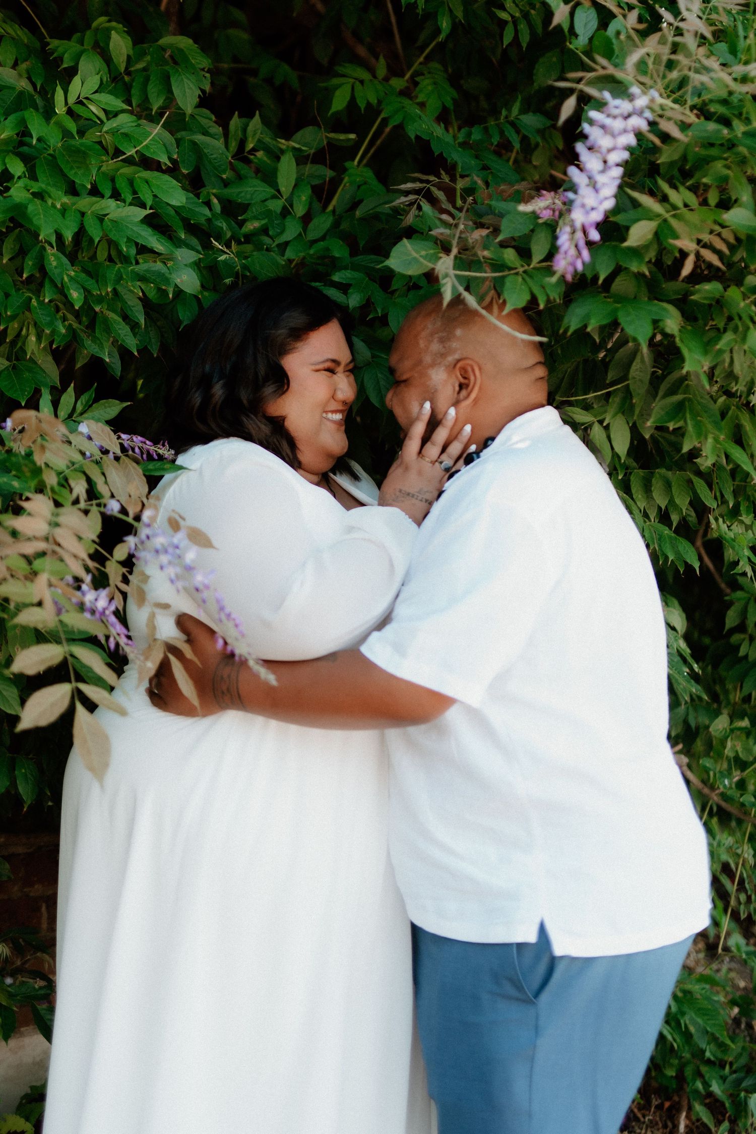 A couple shares a joyful moment together outdoors in matching white outfits against a leafy green backdrop with wisteria.