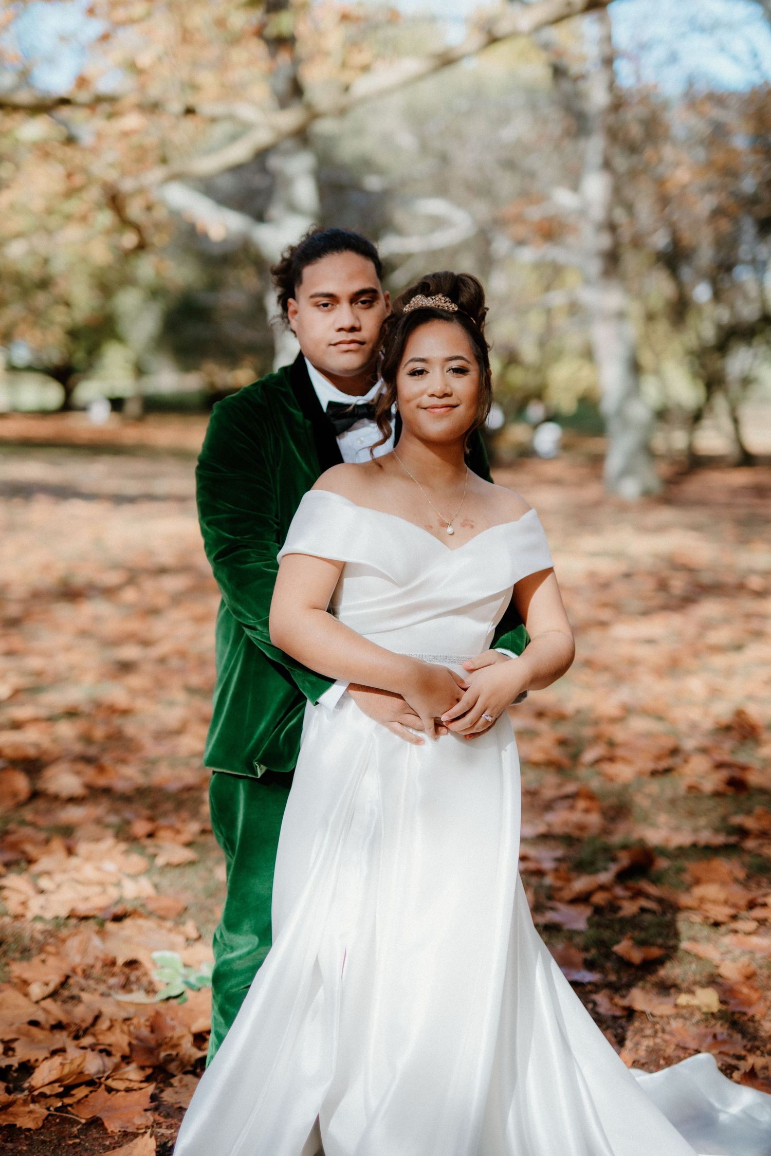 A wedding couple poses together in autumn with fallen leaves, wearing an emerald velvet suit and white off-shoulder gown.