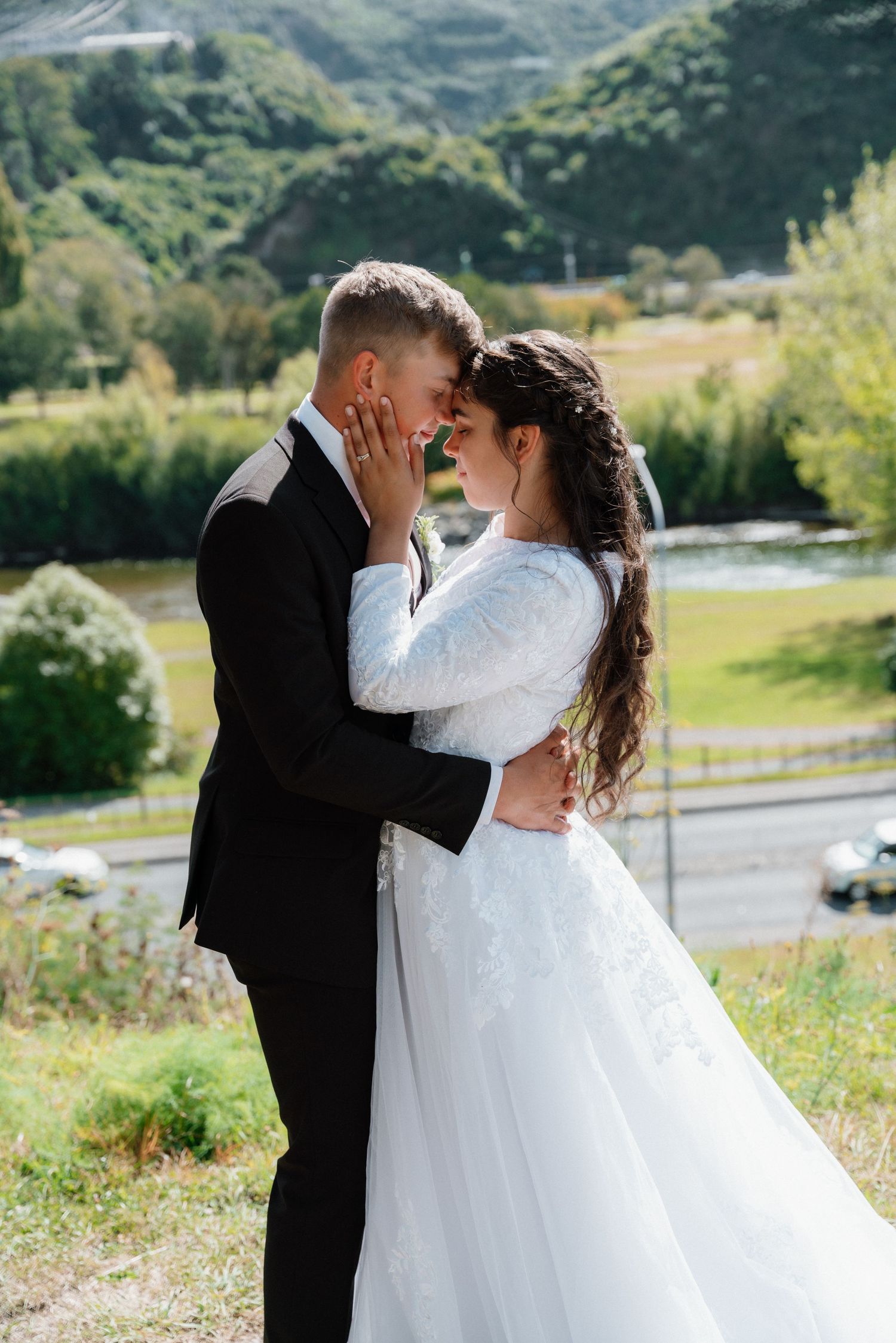 A couple shares an intimate moment in their wedding attire against a scenic mountain backdrop with lush greenery.