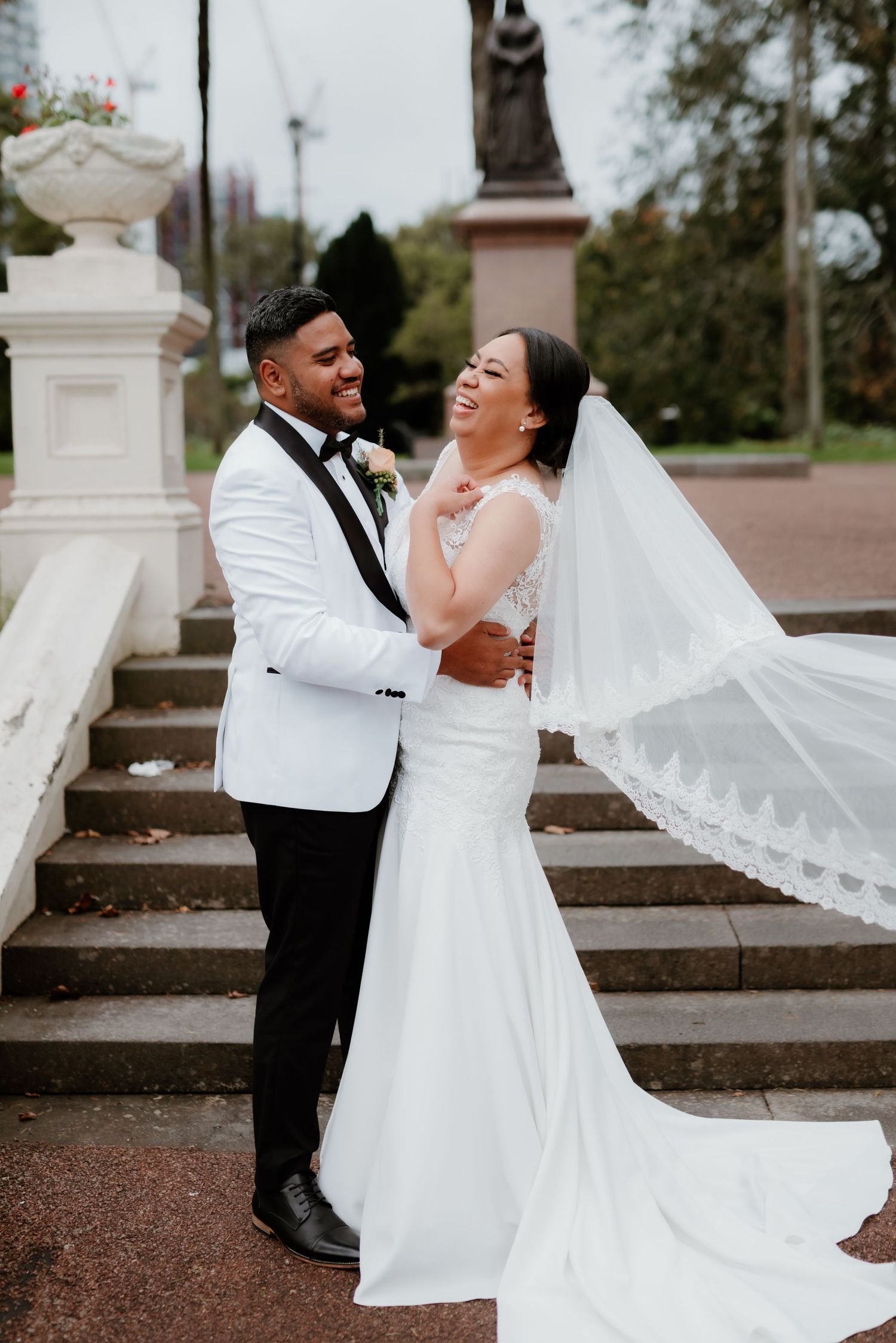 Joyful newlyweds share a candid laughing moment on stone steps, with bride in lace gown and groom in white tuxedo jacket.