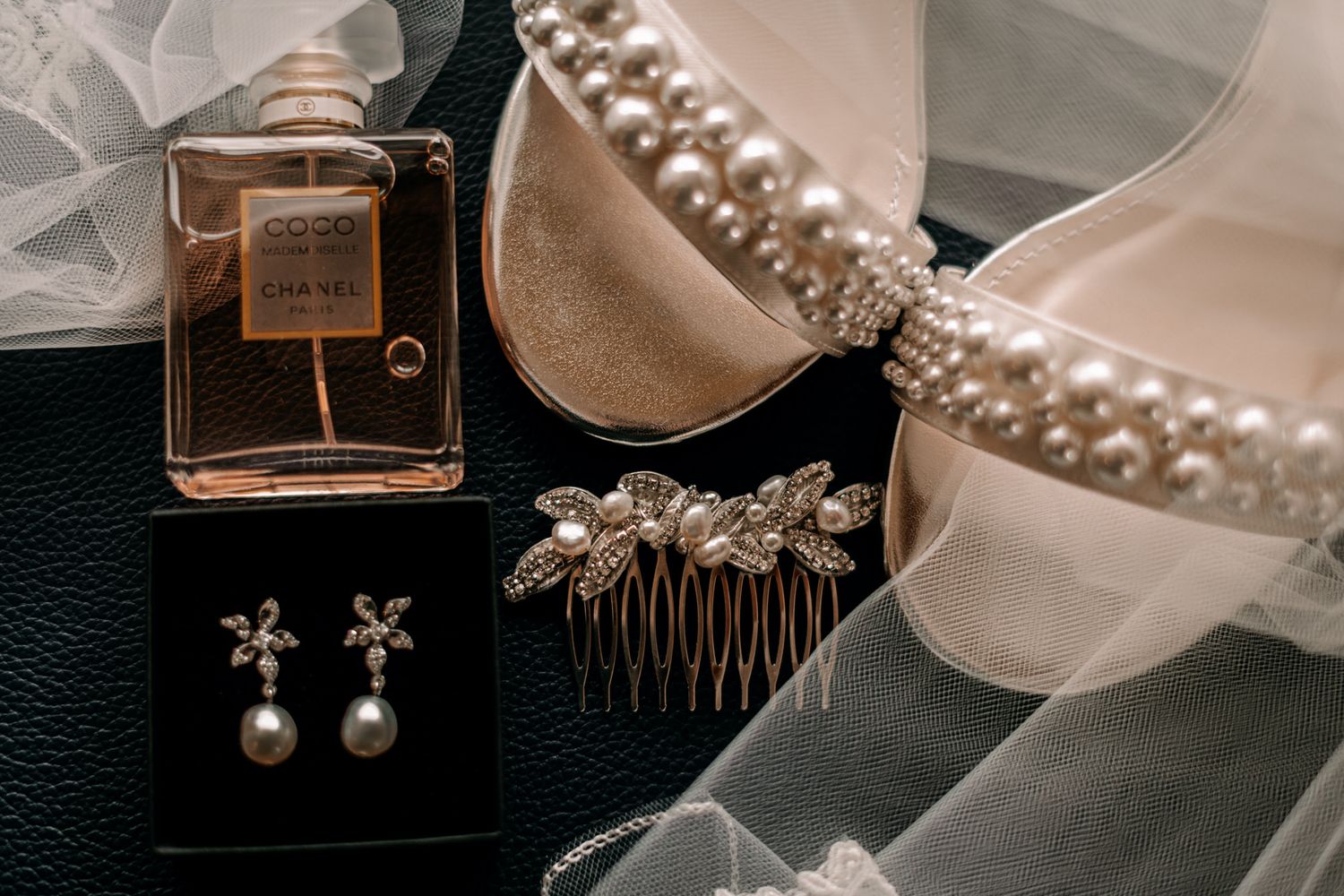 A collection of wedding accessories including pearl necklaces, hair combs, perfume bottles and white ribbons laid out on silk.