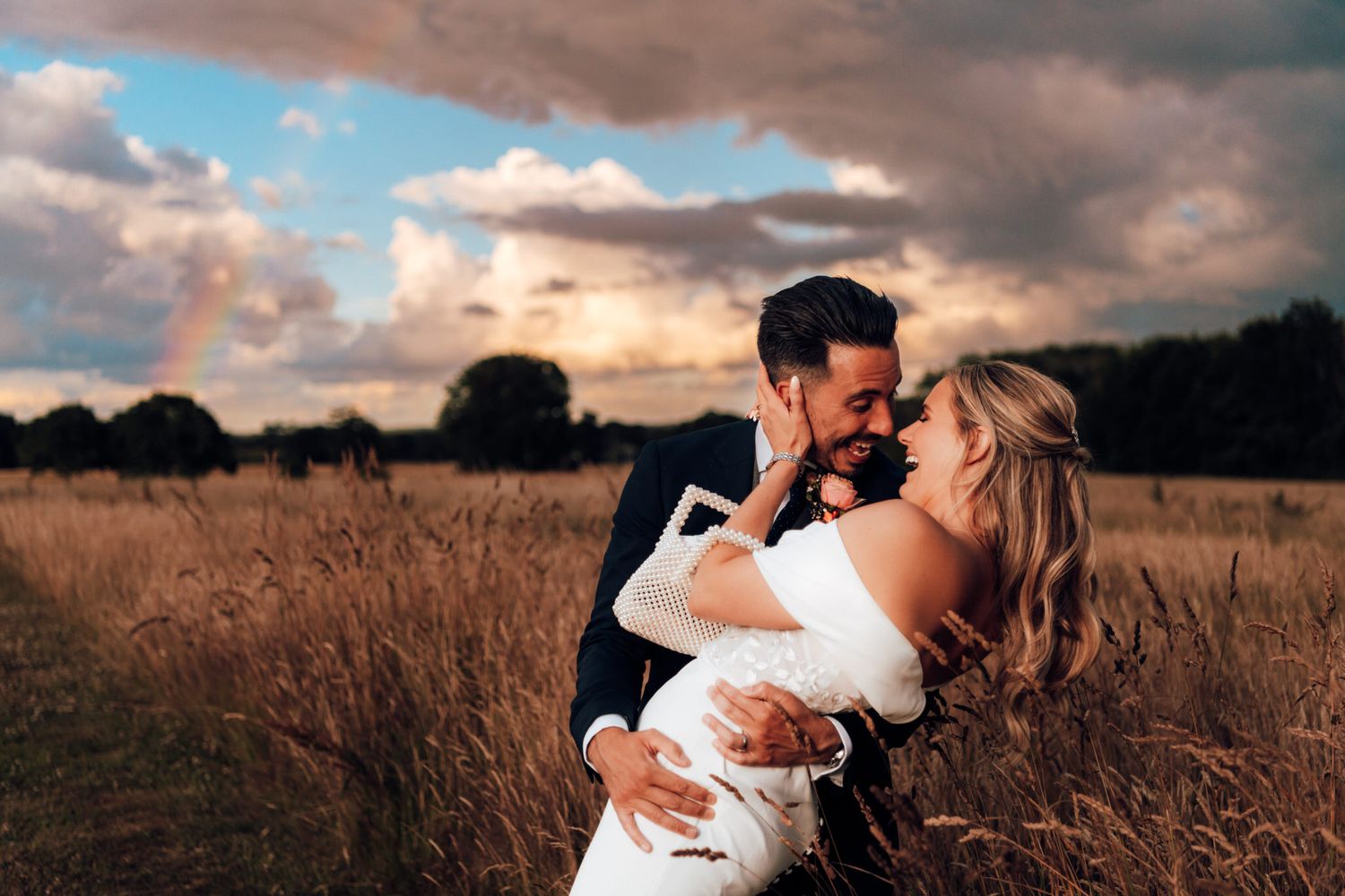 A couple embraces in a golden wheat field at sunset with dramatic clouds in the sky.