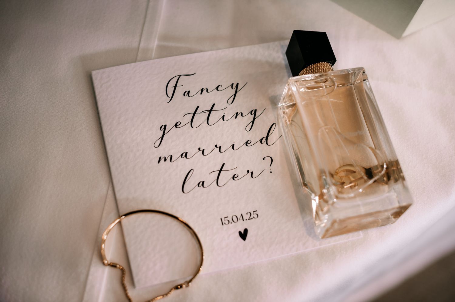 A wedding invitation asking 'Fancy getting married later?' next to a perfume bottle and an elegant golden bracelet.