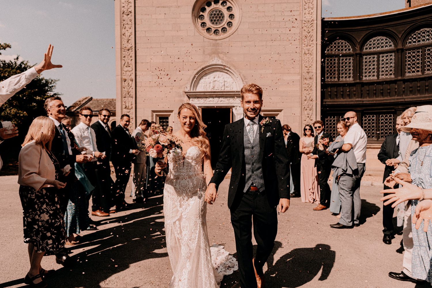 Newlyweds exit church while guests throw confetti in celebration during a summer wedding ceremony.