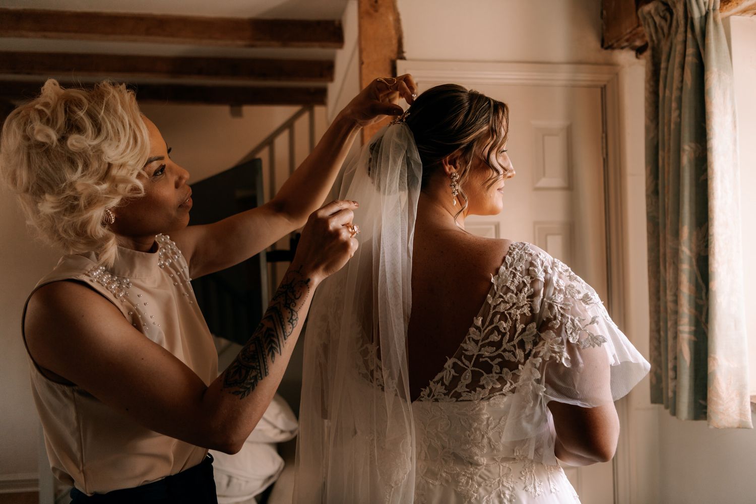 In a rustic room with wooden beams, the soft glow of daylight illuminates a tender wedding preparation moment.
