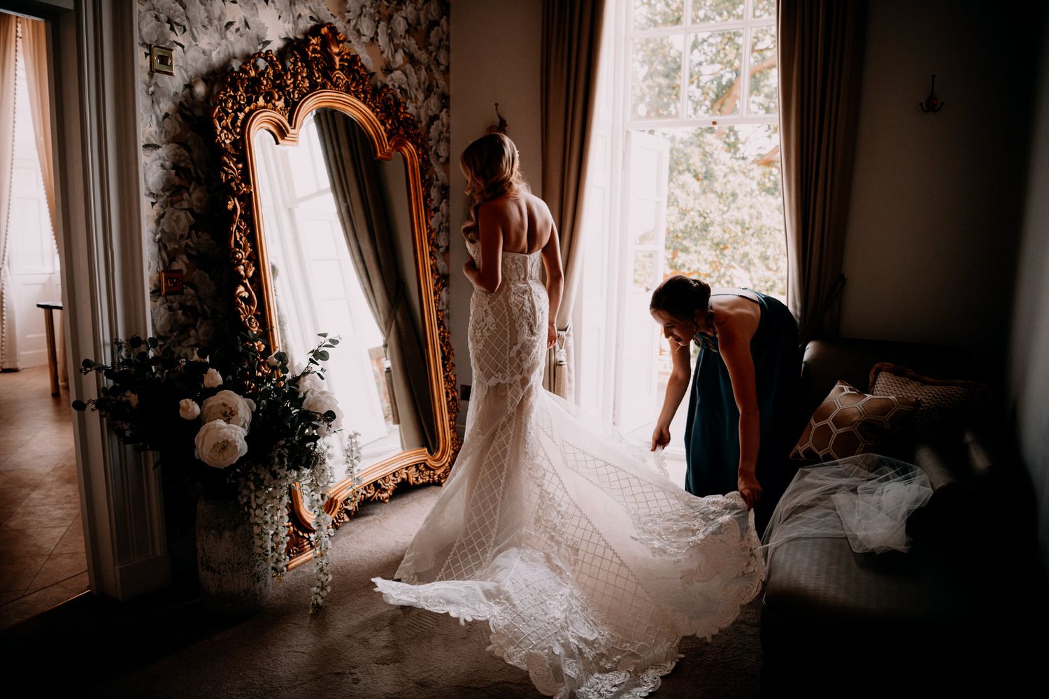 Three figures arrange a flowing white wedding dress train near an ornate gold mirror in an elegant room.