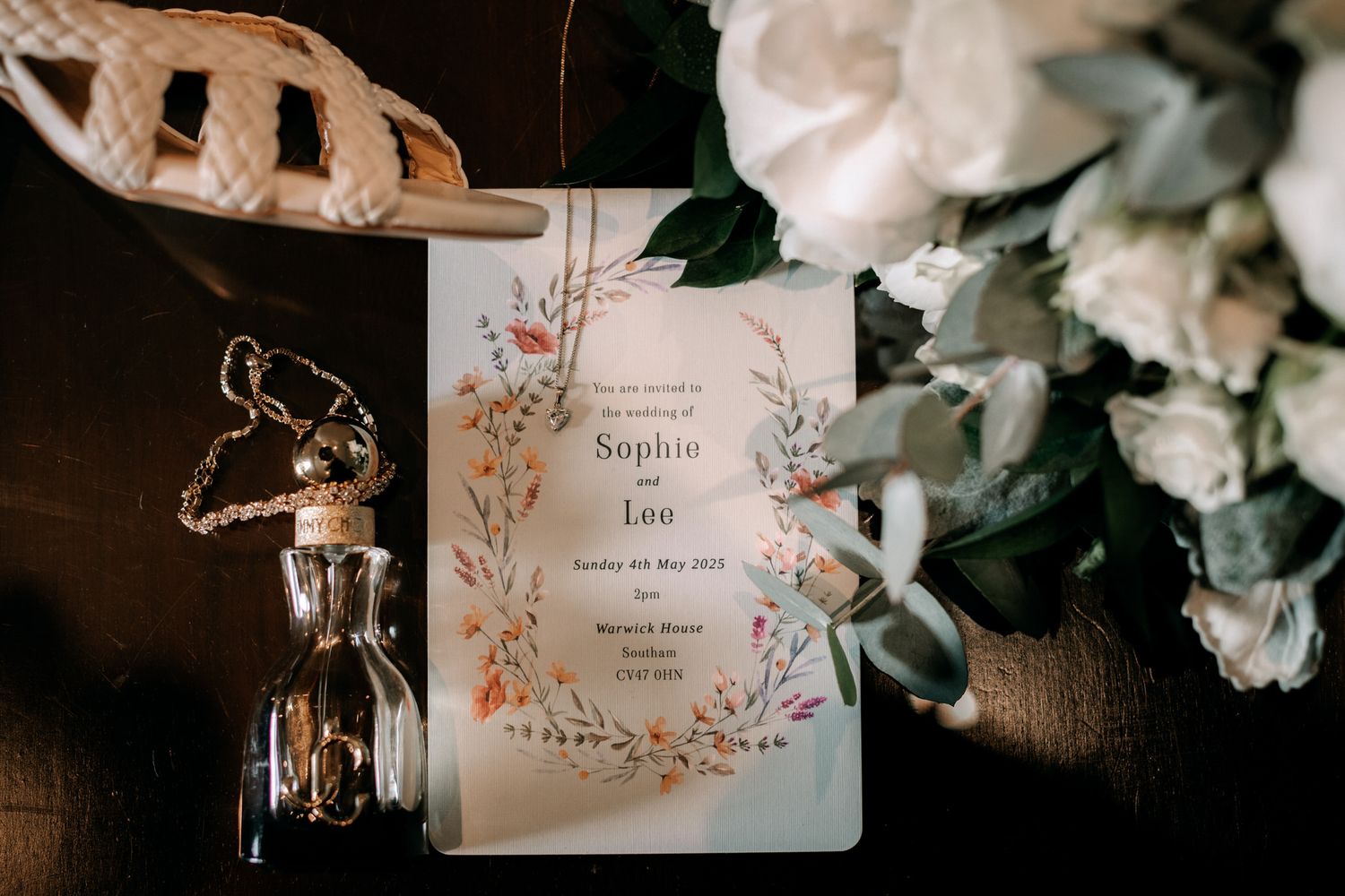 Wedding invitation with floral design lays on dark surface beside white flowers and decorative glass bottle.