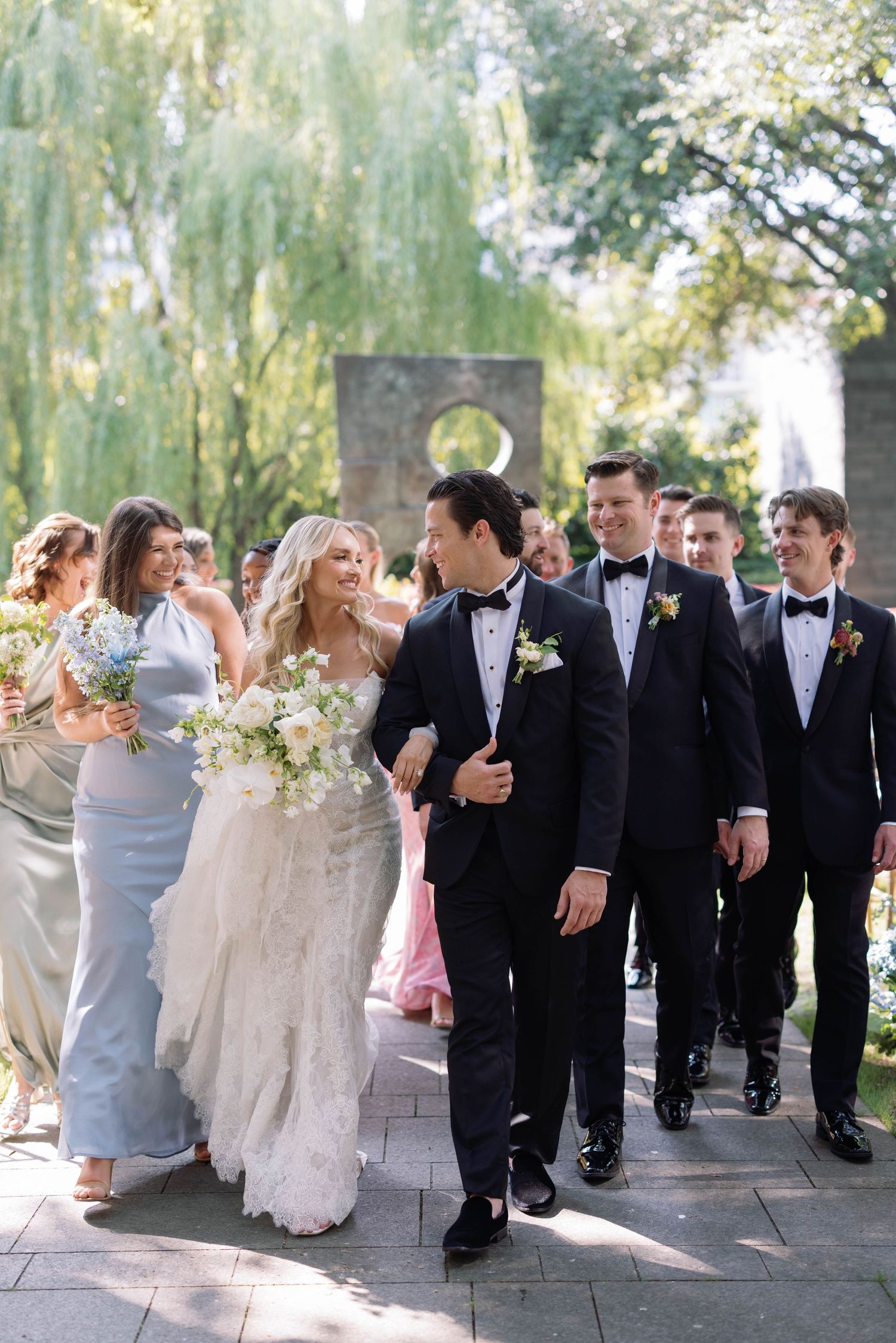 Wedding party walks along garden path with weeping willow trees in background as bridesmaids and groomsmen follow couple.