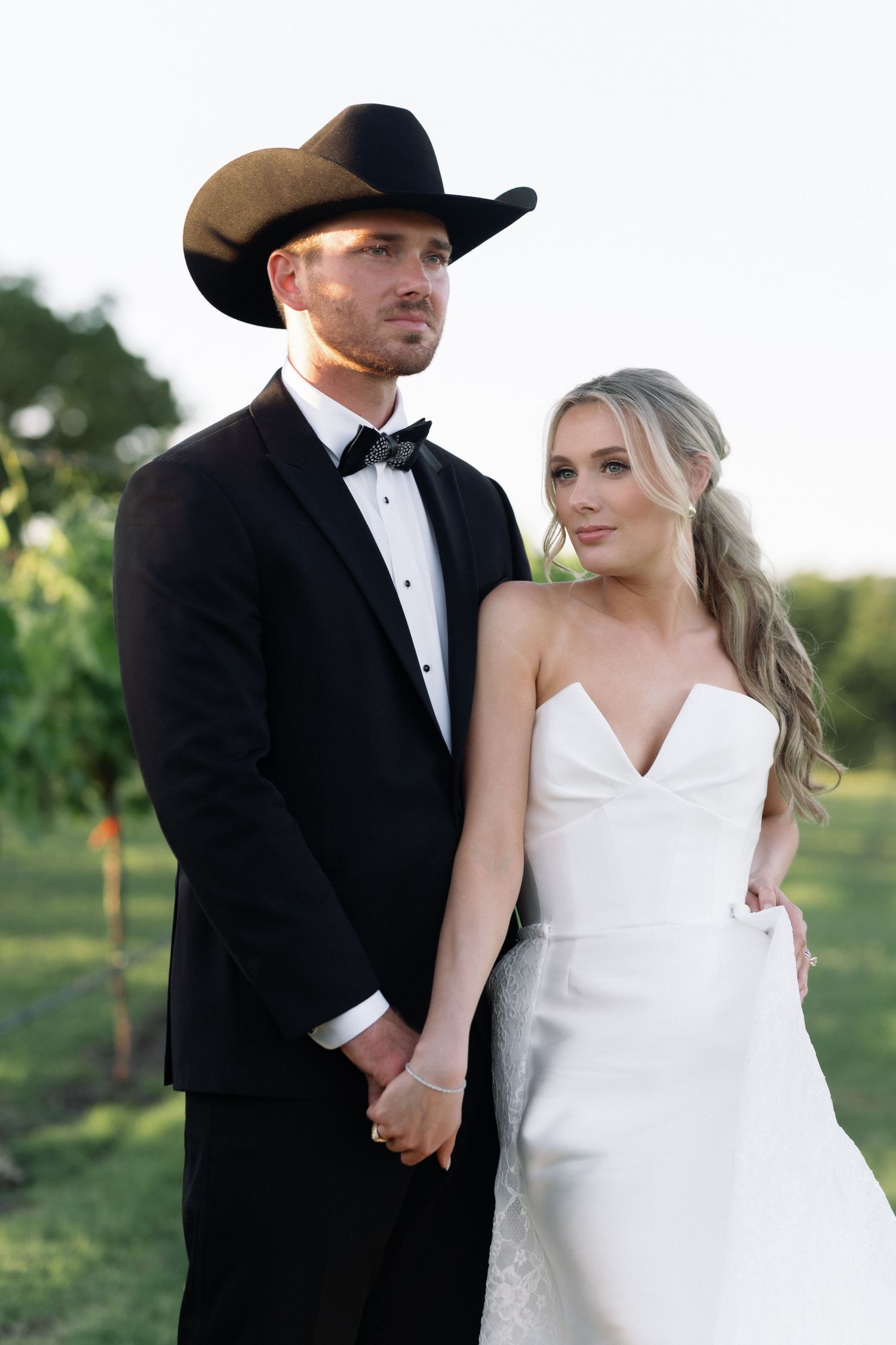 A bride and groom in western wedding attire pose together in a vineyard at sunset.