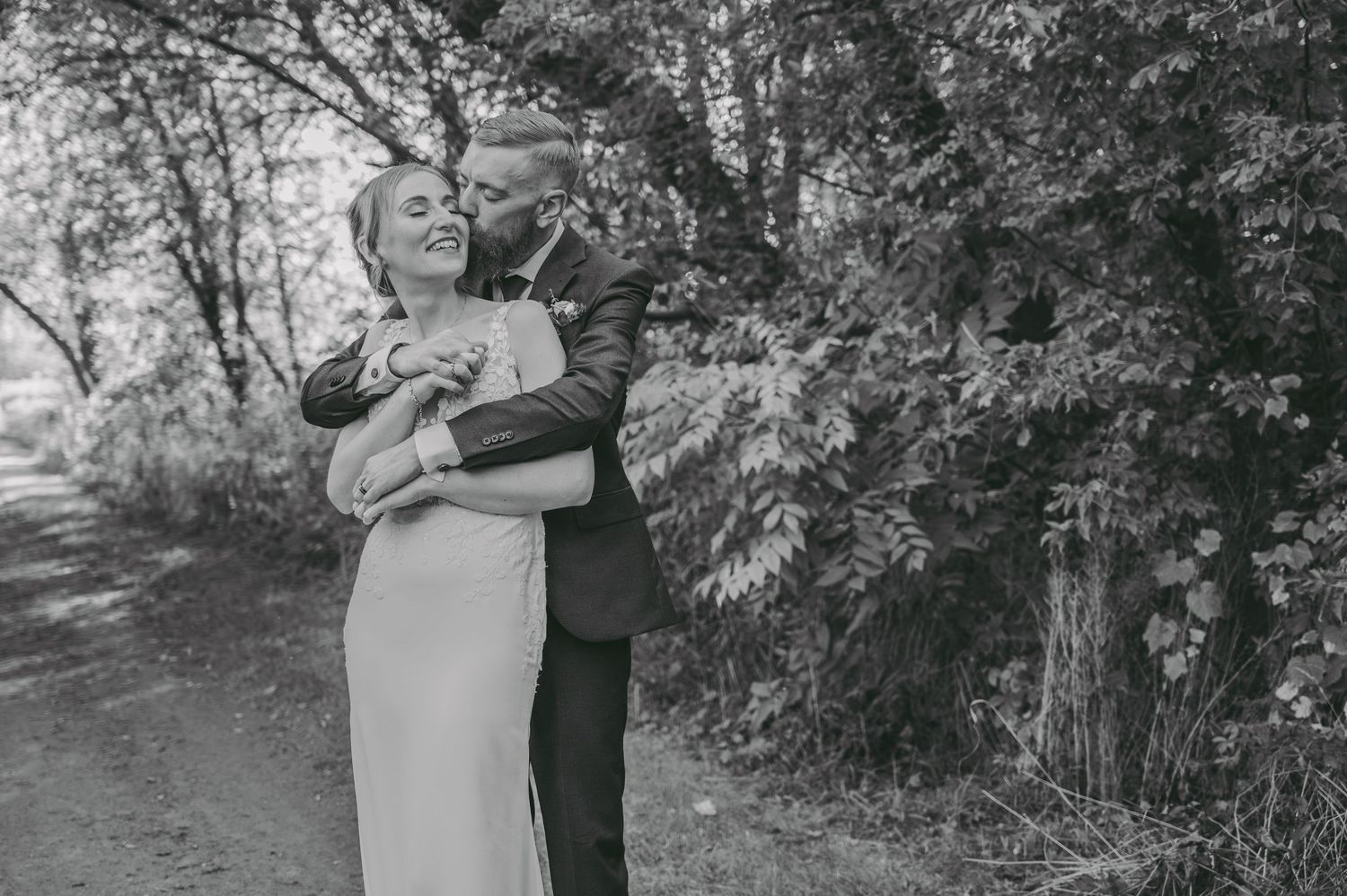 Intimate black and white portrait of a couple embracing outdoors on a wooded path.