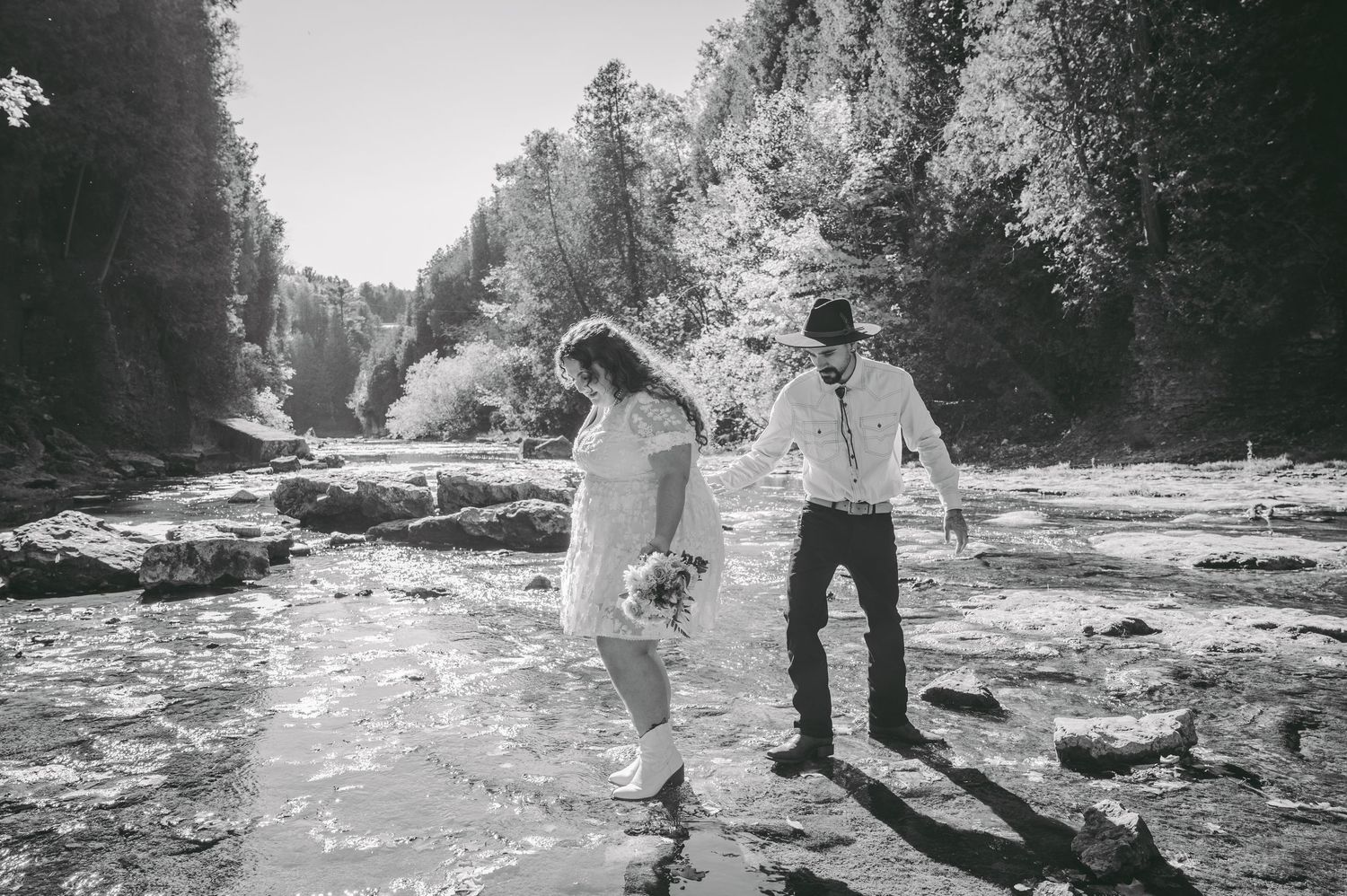 A black and white photo of a couple dancing on rocks by a rushing stream.
