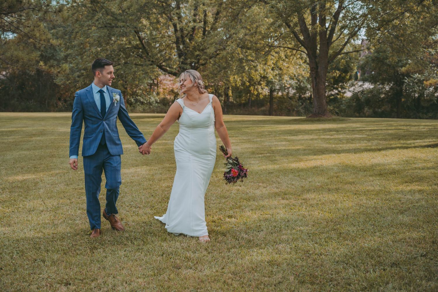 Newlywed couple walking together on grassy hill during autumn wedding photoshoot.