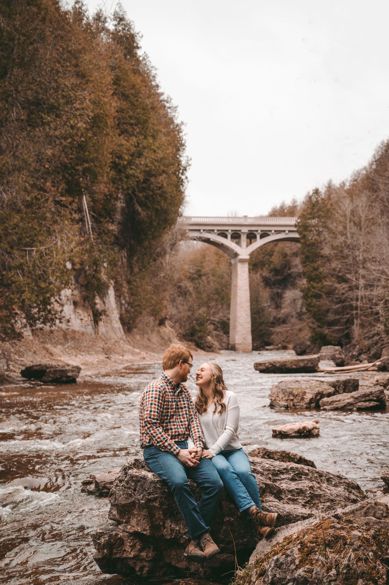 A couple sits on rocky terrain near a bridge.