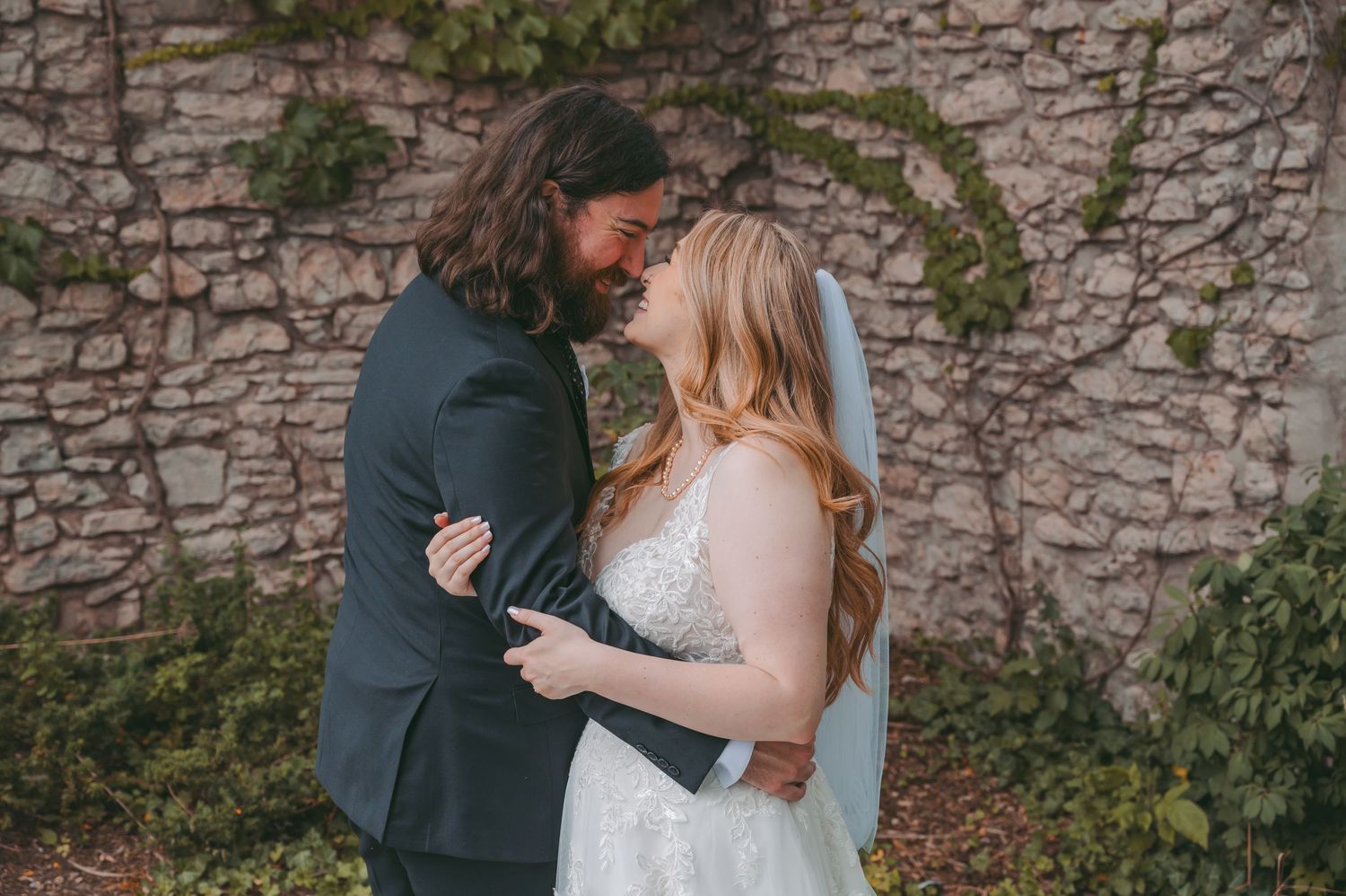 A tender moment between newlyweds against a rustic stone wall covered in ivy.