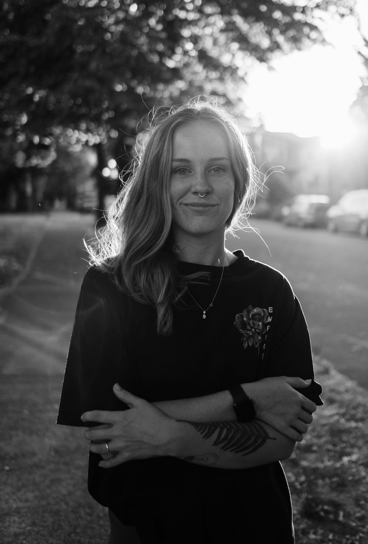 A person in a black sweater stands outdoors in sunlight with arms crossed during golden hour.
