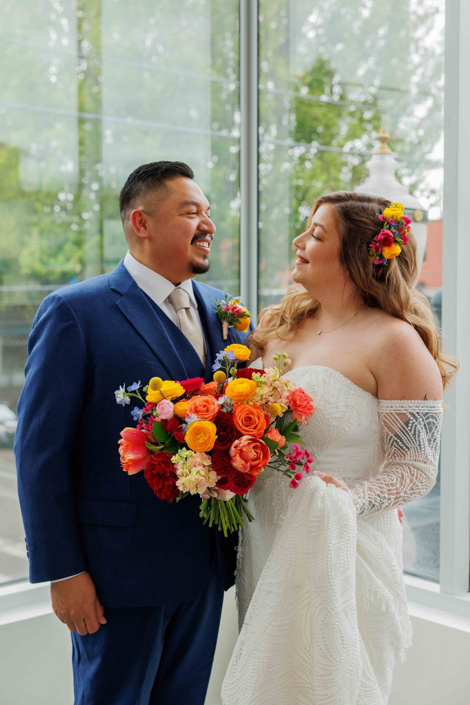 Wedding couple shares joyful laughter in bright sunlit venue with vibrant orange and red floral bouquet accents.
