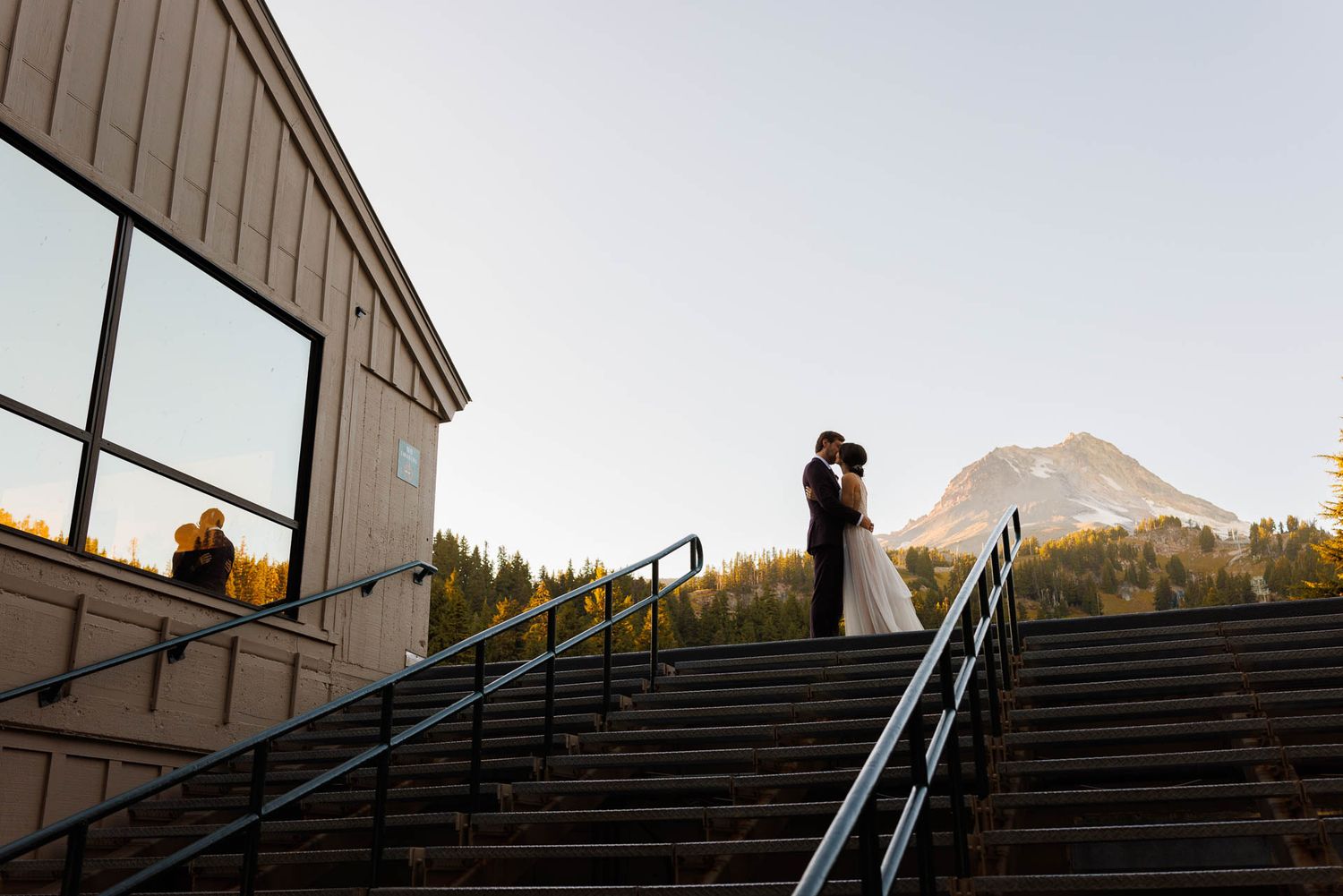 A silhouetted couple embraces on steps outside a mountain venue at sunset.