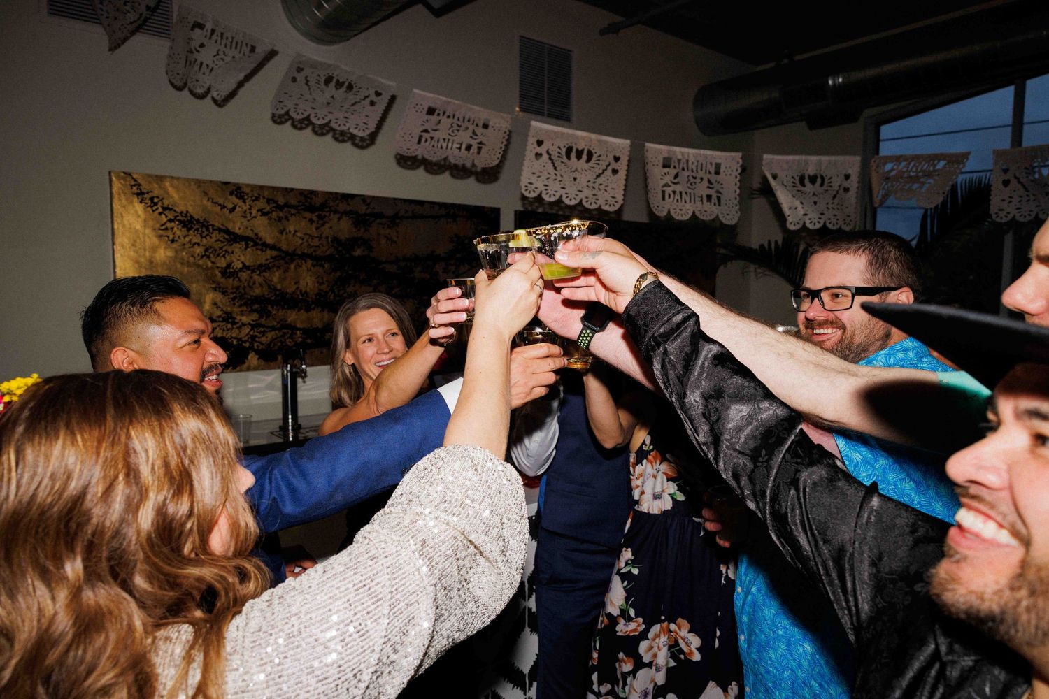 Wedding reception guests raising glasses in a celebratory toast during a dimly lit party.