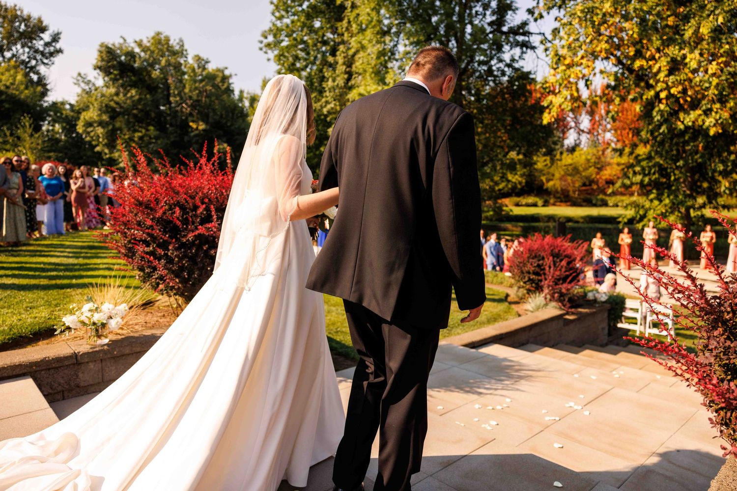 Bride in flowing white gown walking down outdoor aisle with vibrant red autumn landscaping.