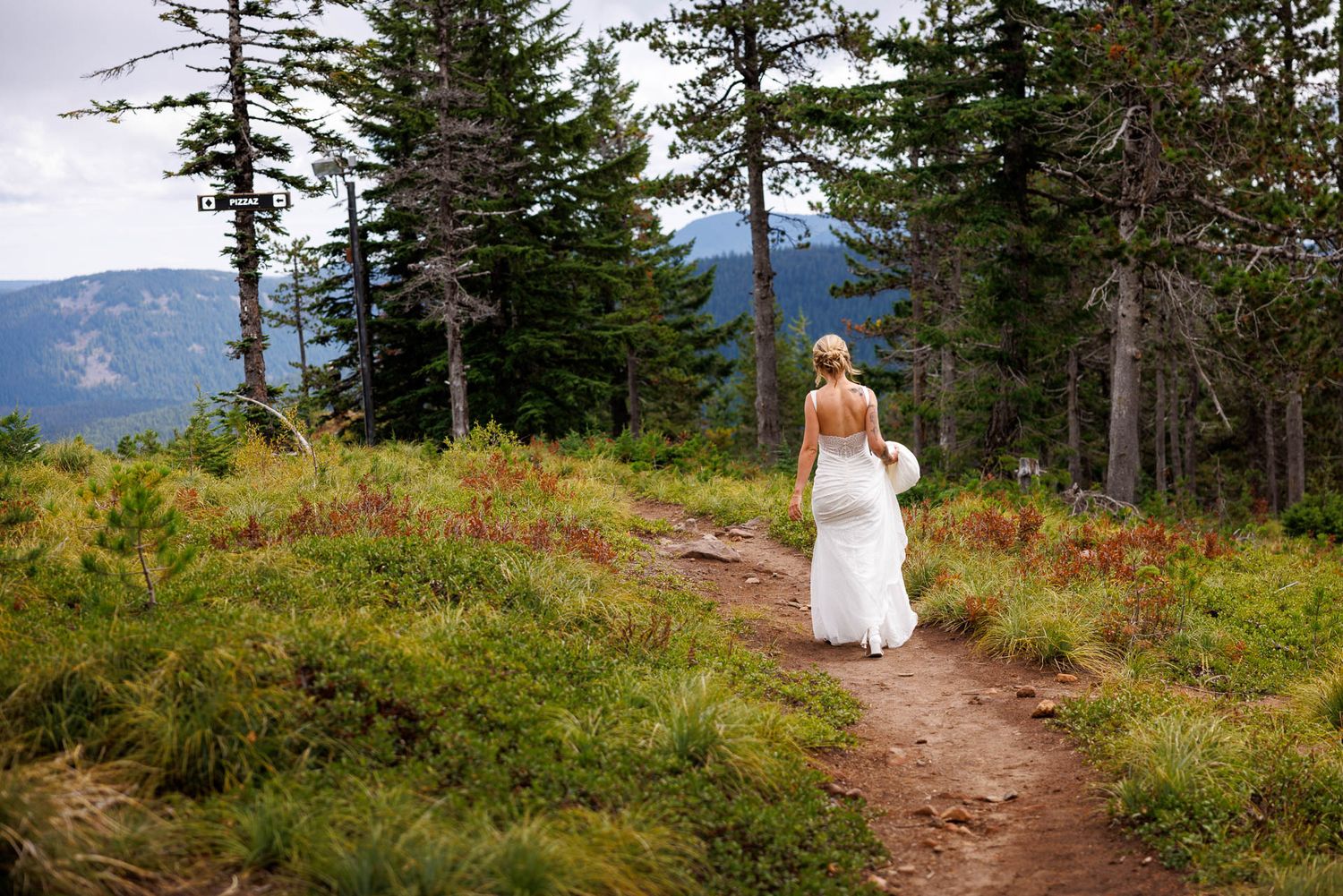 Person in flowing white wedding dress walks along mountain trail surrounded by evergreen forest.